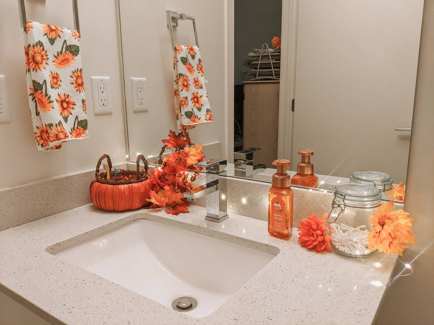 A small bathroom vanity with a pumpkin soap dispenser, sunflower towels, and string lights.