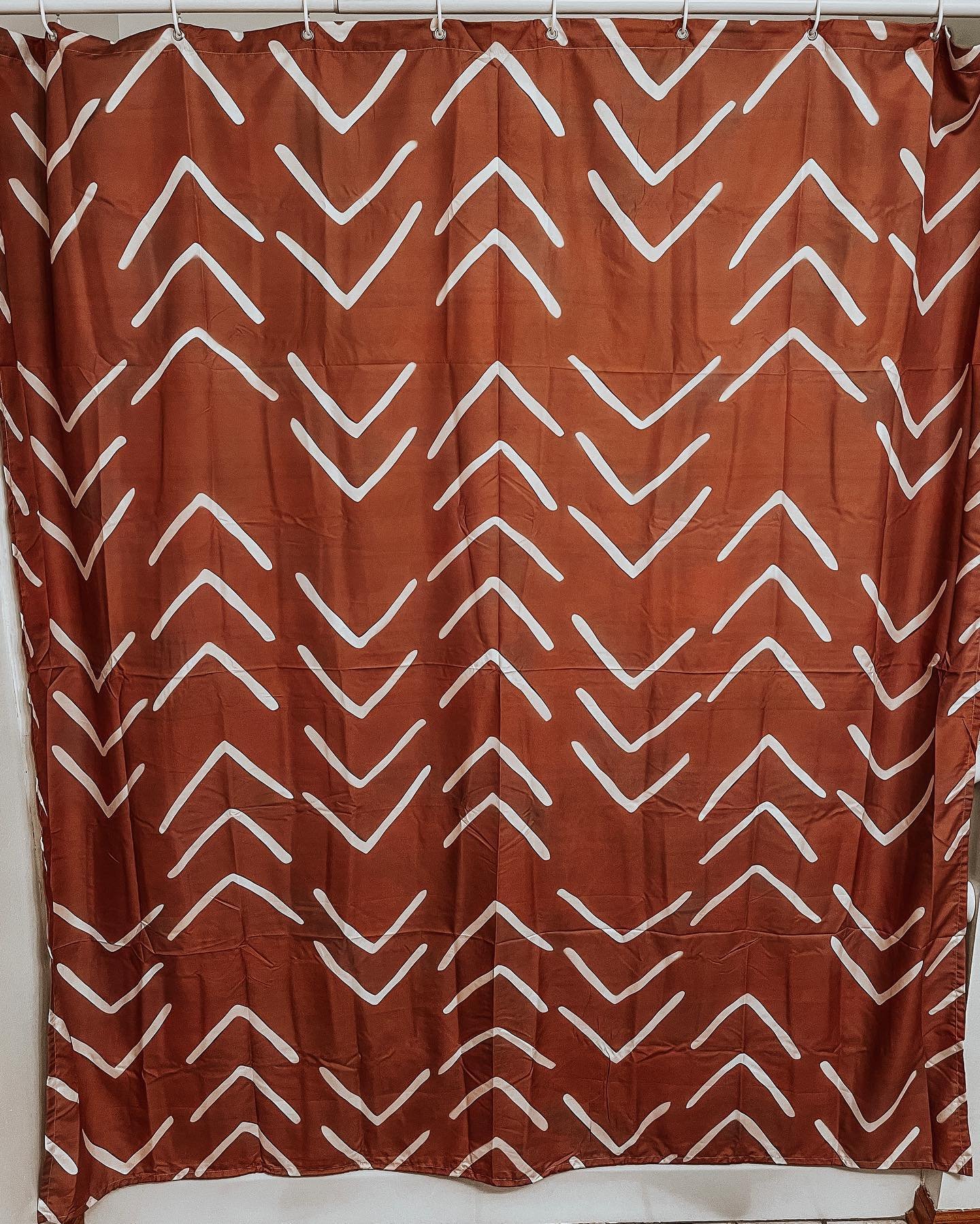 A deep terracotta orange/brown shower curtain featuring an all-over repeating pattern of hand-drawn white chevron or arrow symbols.