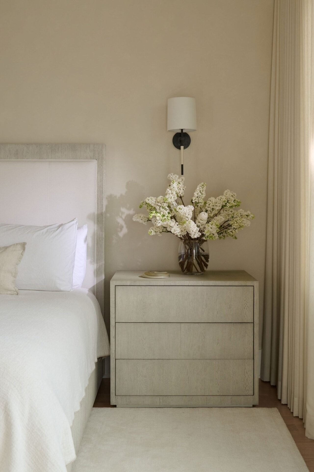 A neutral-toned, textured three-drawer nightstand with no visible pulls. The top holds a large, clear vase filled with white flowers. A modern black and white wall sconce is mounted above the nightstand.
