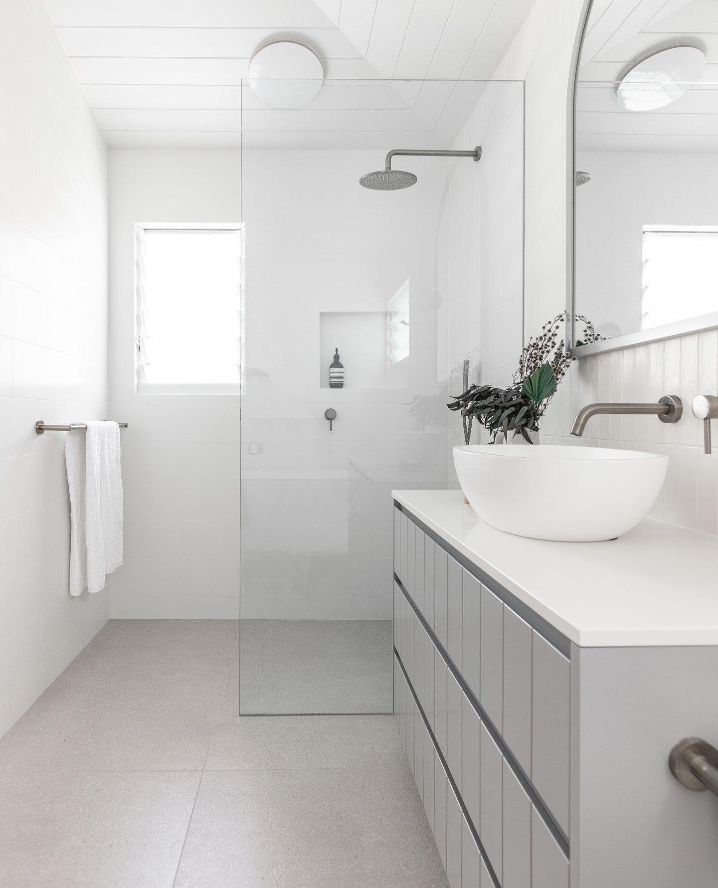 A bright, all-white wet room shower with a single frameless glass panel, gunmetal gray fixtures, and a light gray tiled floor.