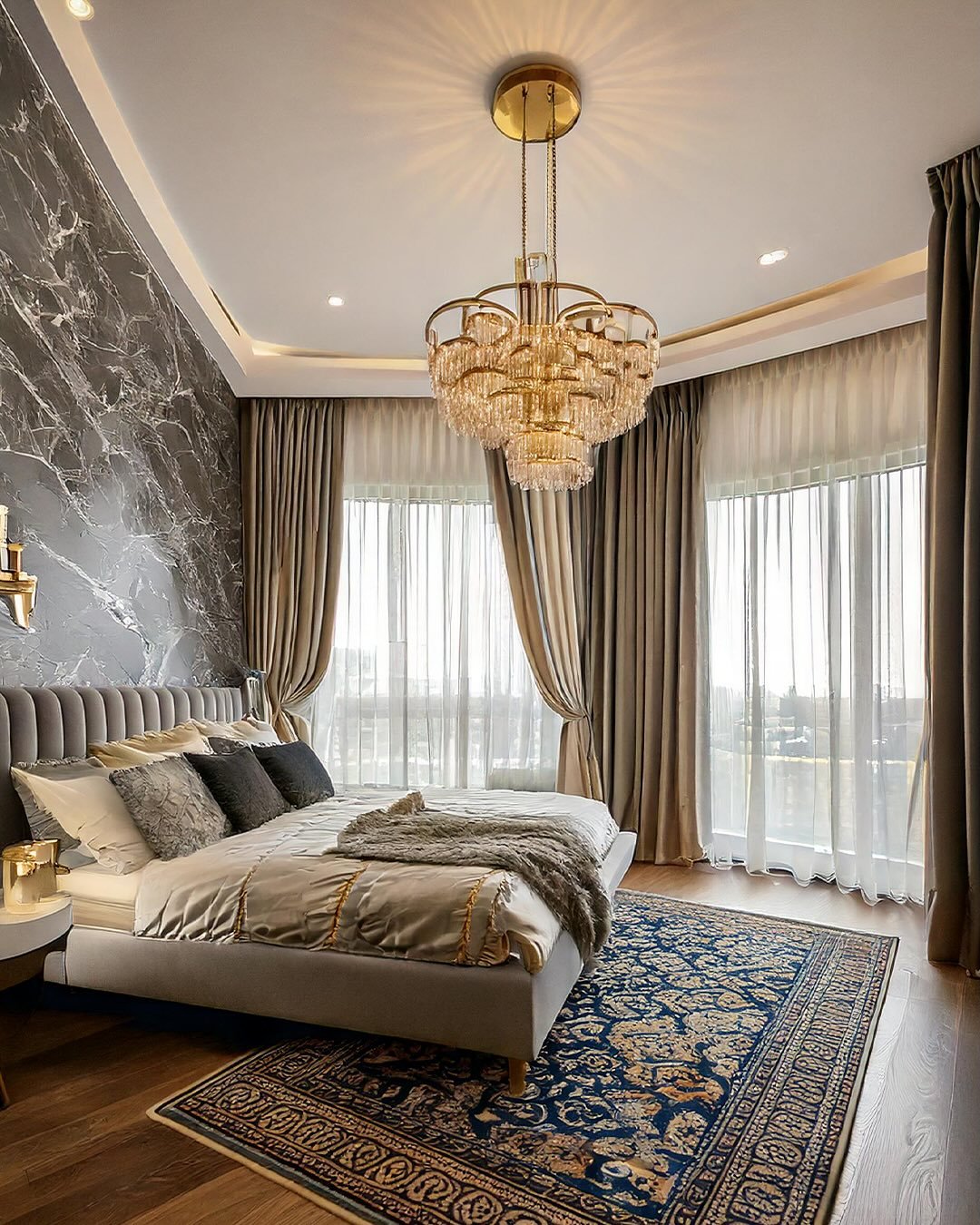A luxurious, multi-tiered gold-toned chandelier with clear crystal rods hangs in a high-end bedroom with dark marble walls and floor-to-ceiling windows.