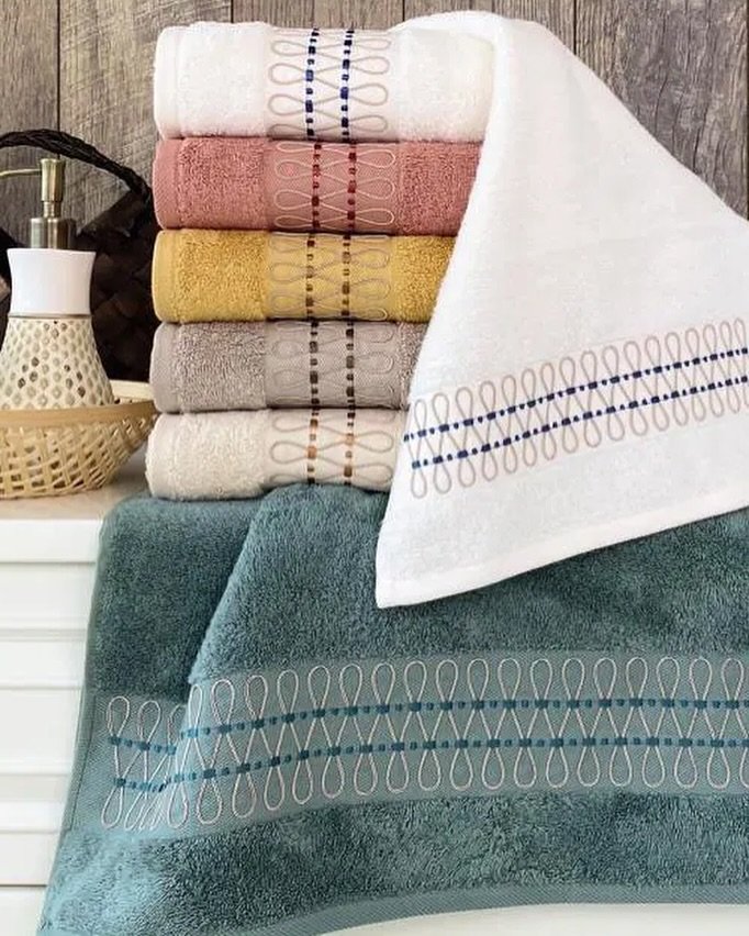Stack of six colorful bath towels with a repetitive looped embroidery pattern, with a teal towel draped in front.
