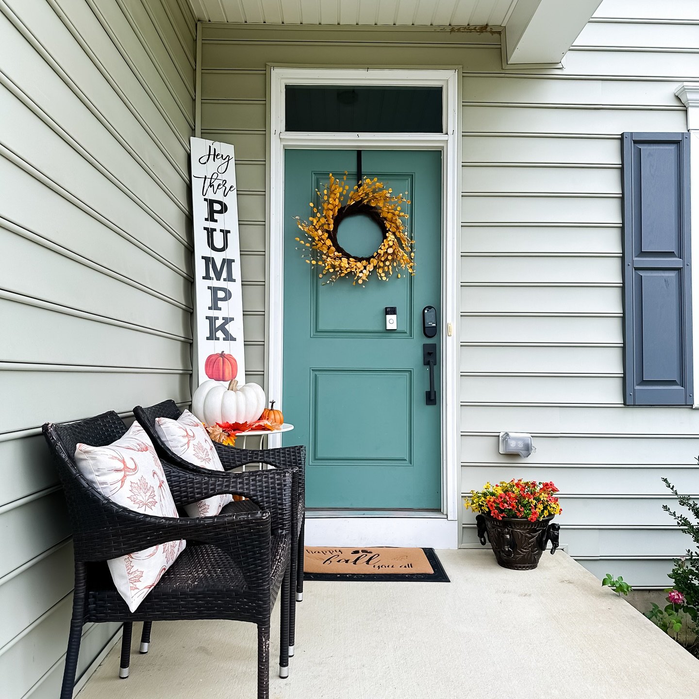 A cozy front porch with a teal door, decorated for fall with a yellow berry wreath, a white vertical sign, and a small sitting area with pillows.