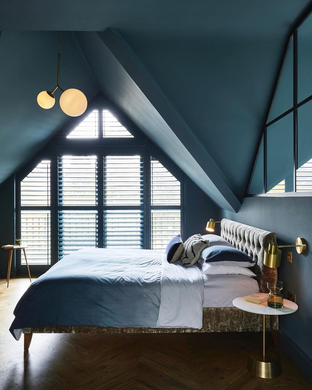 A dramatic loft bedroom painted in a deep blue, with a large triangular window, a tufted headboard, and minimalist modern light fixtures.