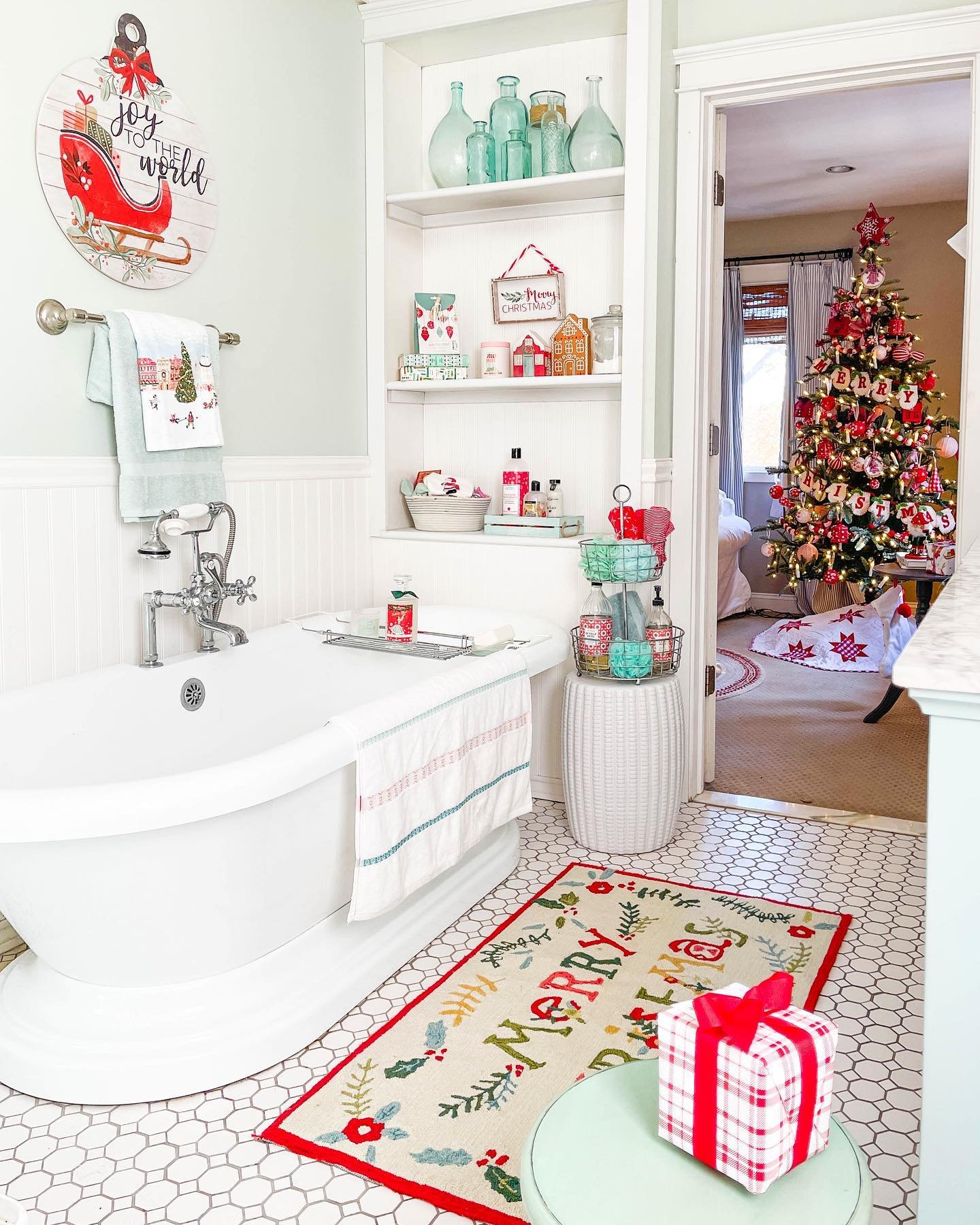 A mint green bathroom with a clawfoot tub, a festive "MERRY & BRIGHT" floor rug, and a large, decorated Christmas tree visible through the open doorway.