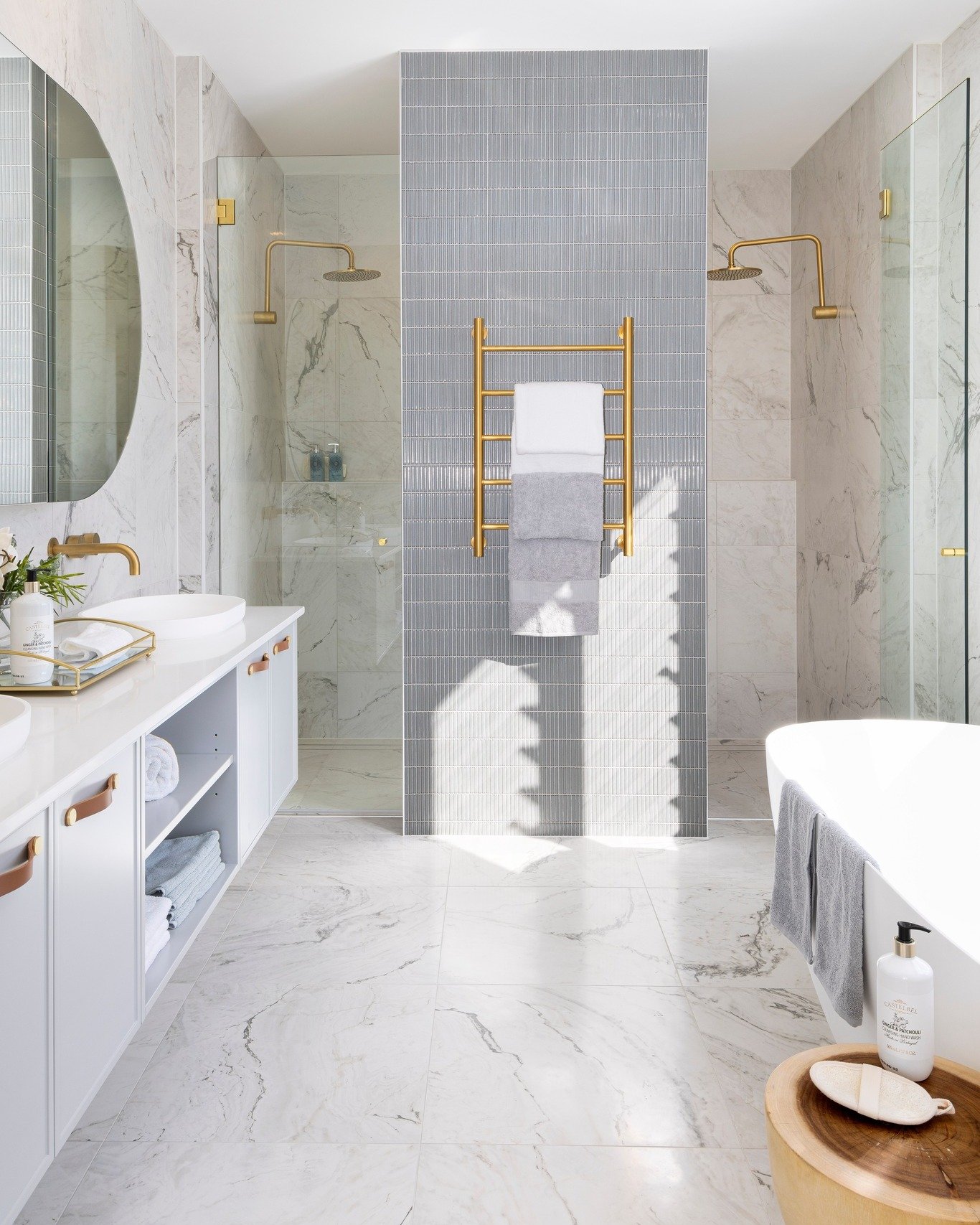 A luxurious bathroom with a double shower, featuring a central partition with vertical grey tiles, white marble tiles, and gold fixtures.