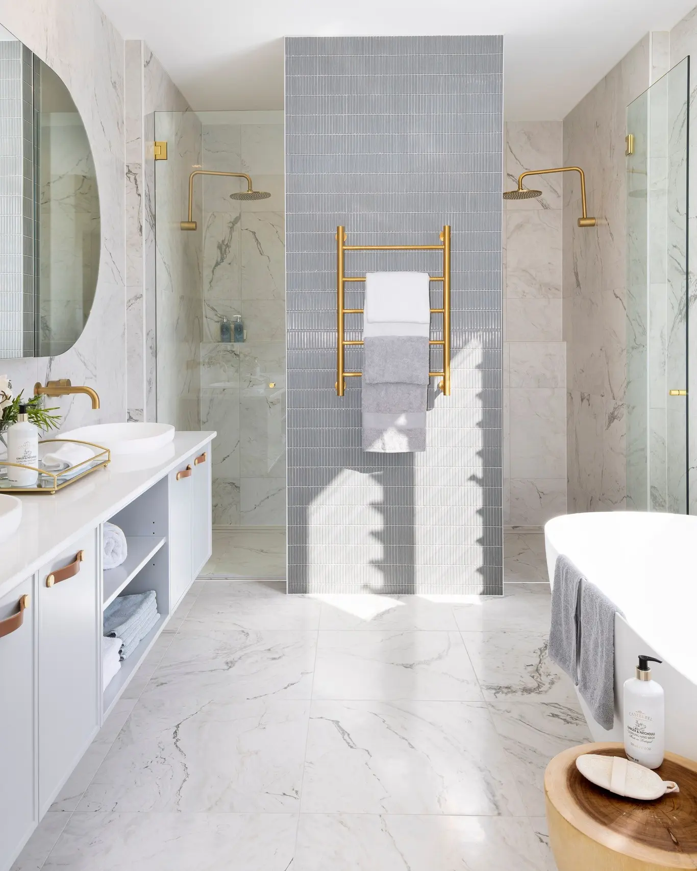 A luxurious bathroom with a double shower, featuring a central partition with vertical grey tiles, white marble tiles, and gold fixtures.