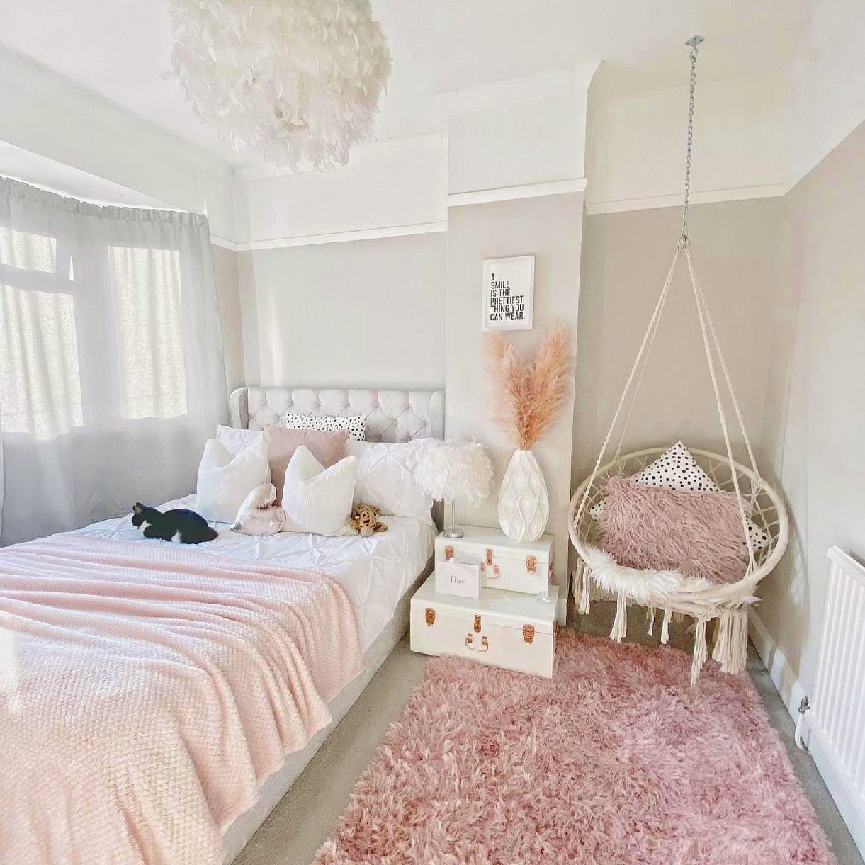 A soft and luxurious bedroom with a tufted white bed, a pink shaggy rug, a white feather chandelier, and a hanging macrame chair with a fluffy pink pillow.
