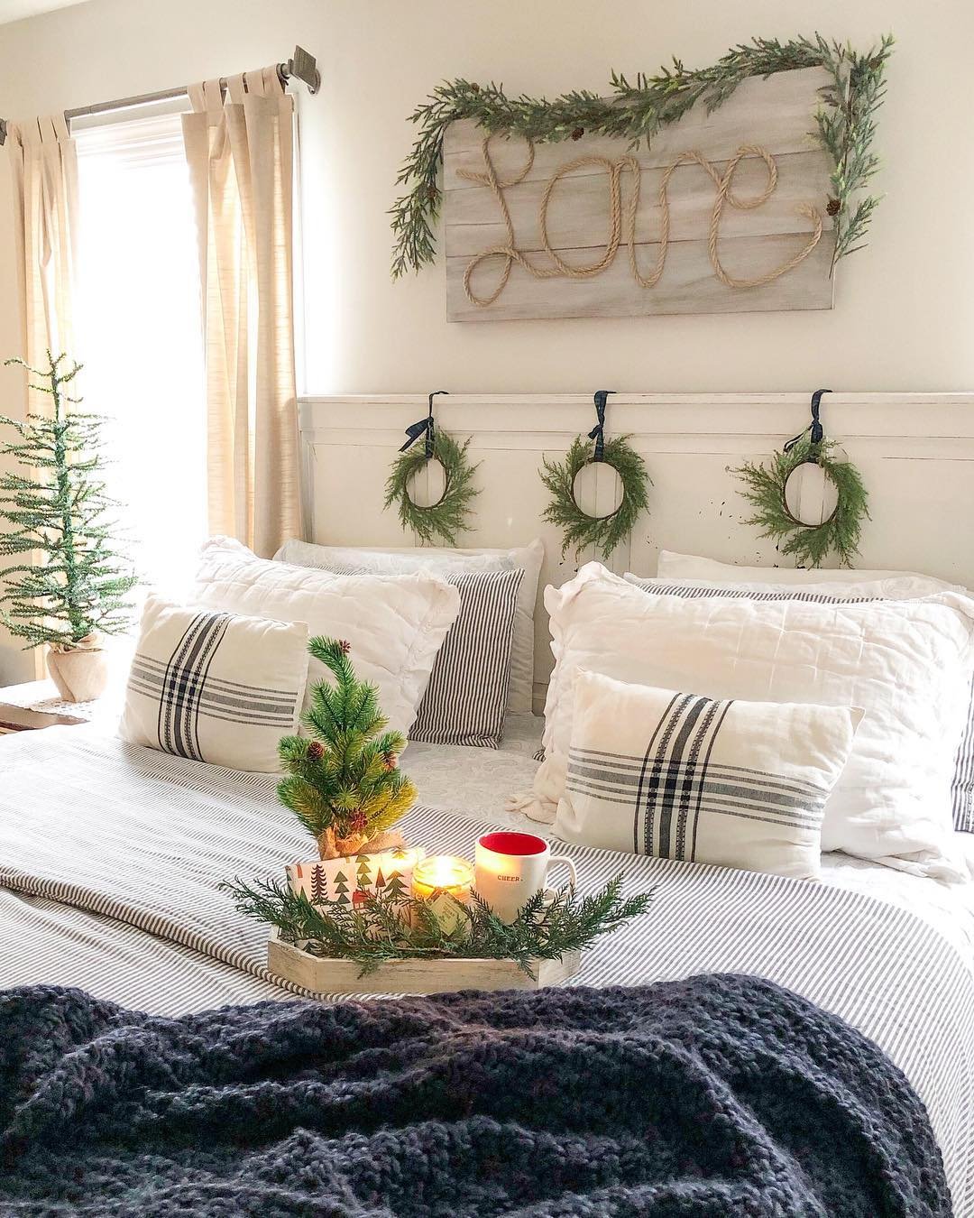A cozy farmhouse bedroom with a white bed, striped bedding, and a distressed wood sign reading "Love" made of rope, decorated with a garland and three mini wreaths.