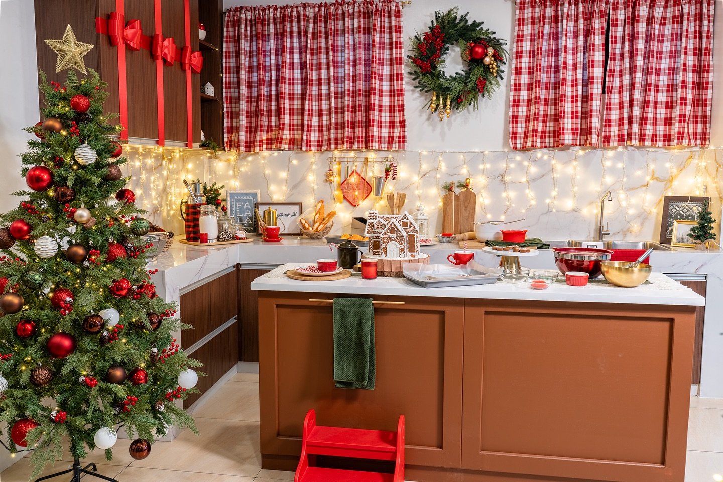 A cozy kitchen fully decorated in red, white, and green for Christmas, featuring a brightly lit Christmas tree, plaid curtains, a large wreath, and a central island set up as a baking station with a completed gingerbread house.