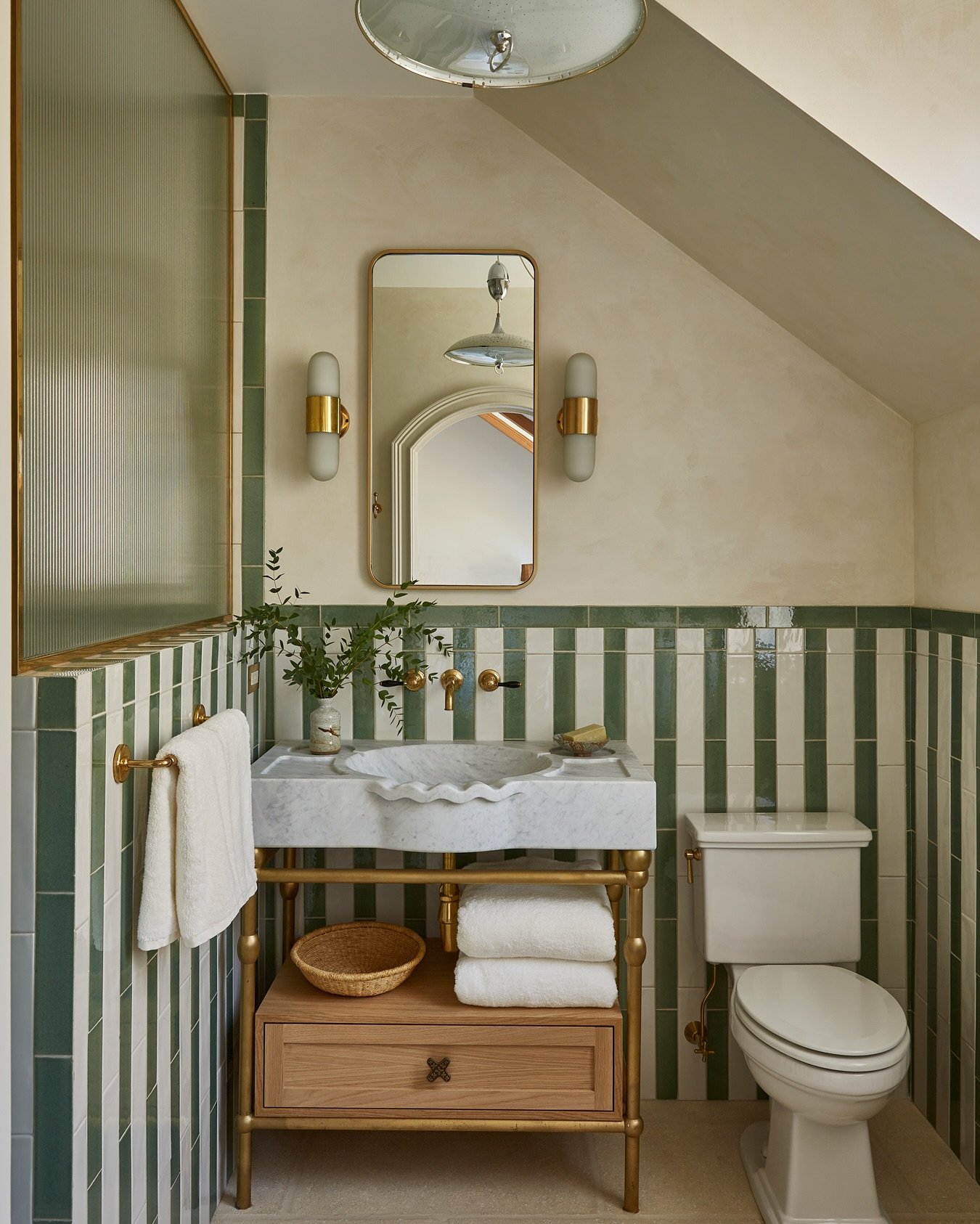 Bathroom with green and white striped tiles and a brass washstand vanity. The vanity features a marble top, a single wooden drawer, and an open lower shelf with white folded towels.