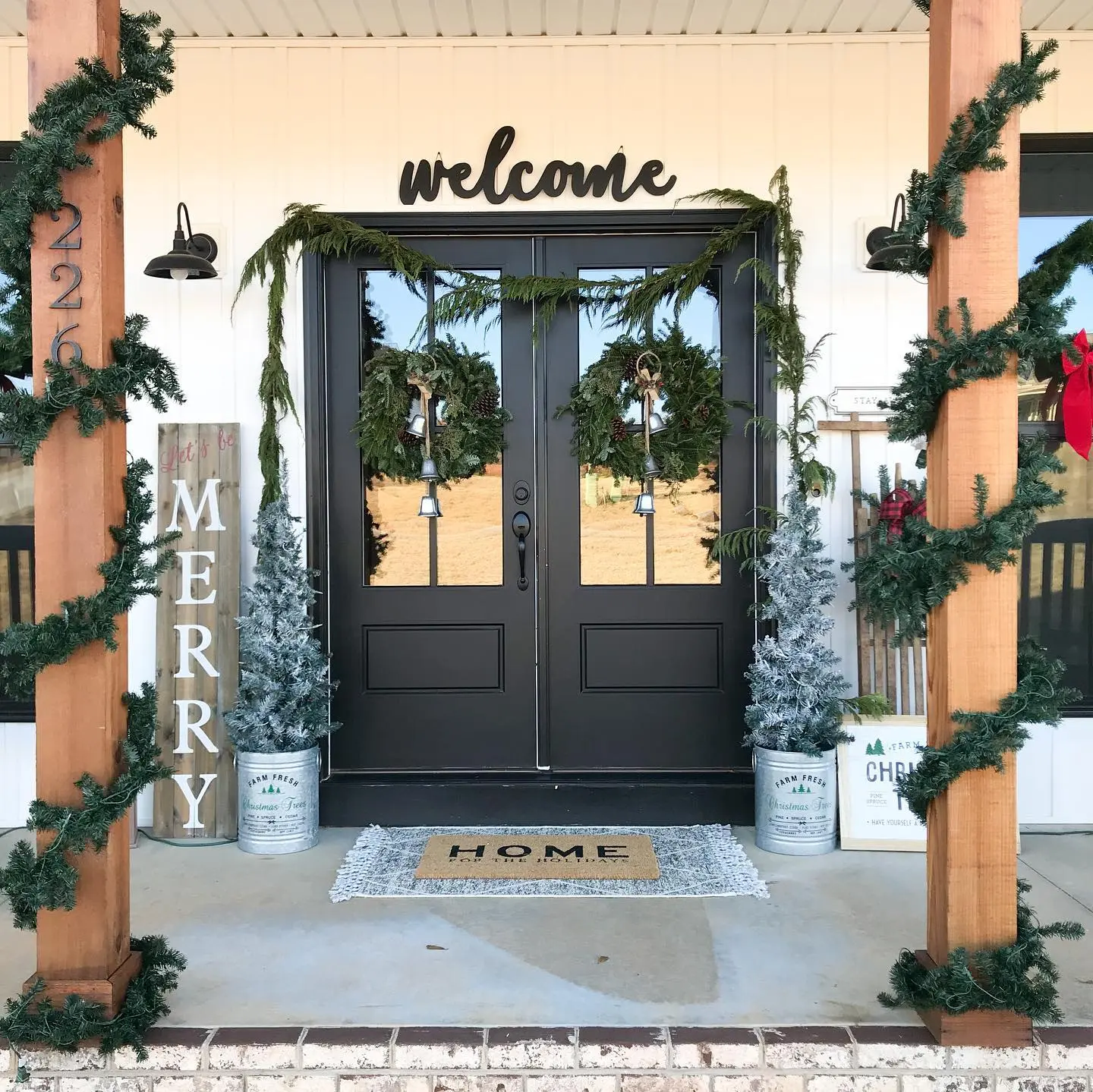 A contemporary farmhouse Christmas porch with exposed wood pillars wrapped in simple garland, black double doors with twin wreaths, and a vertical "MERRY" sign next to frosted mini-trees in galvanized buckets.