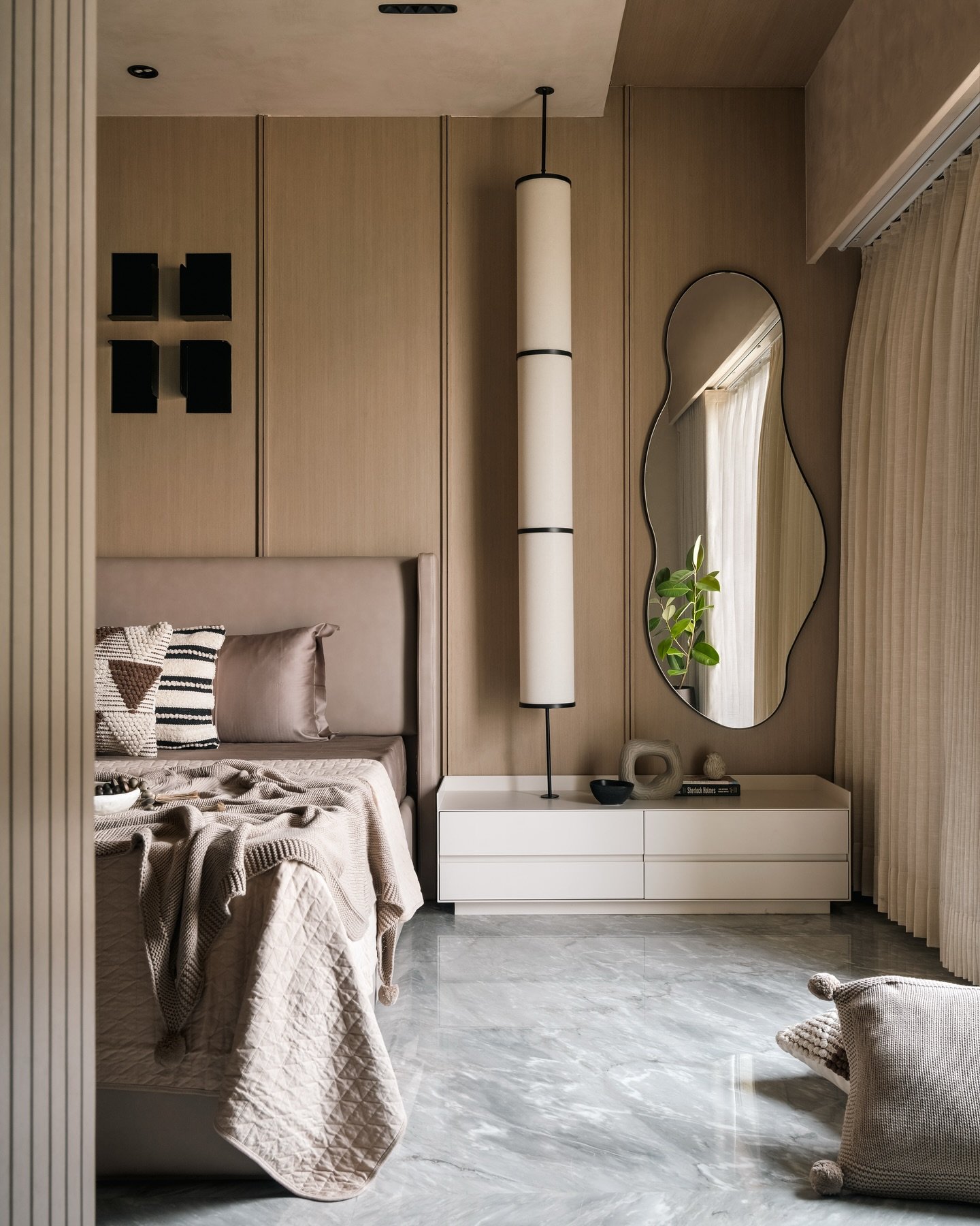 A large, black-framed mirror with an irregular, wavy shape hangs on a wall in a modern, neutral-toned bedroom.