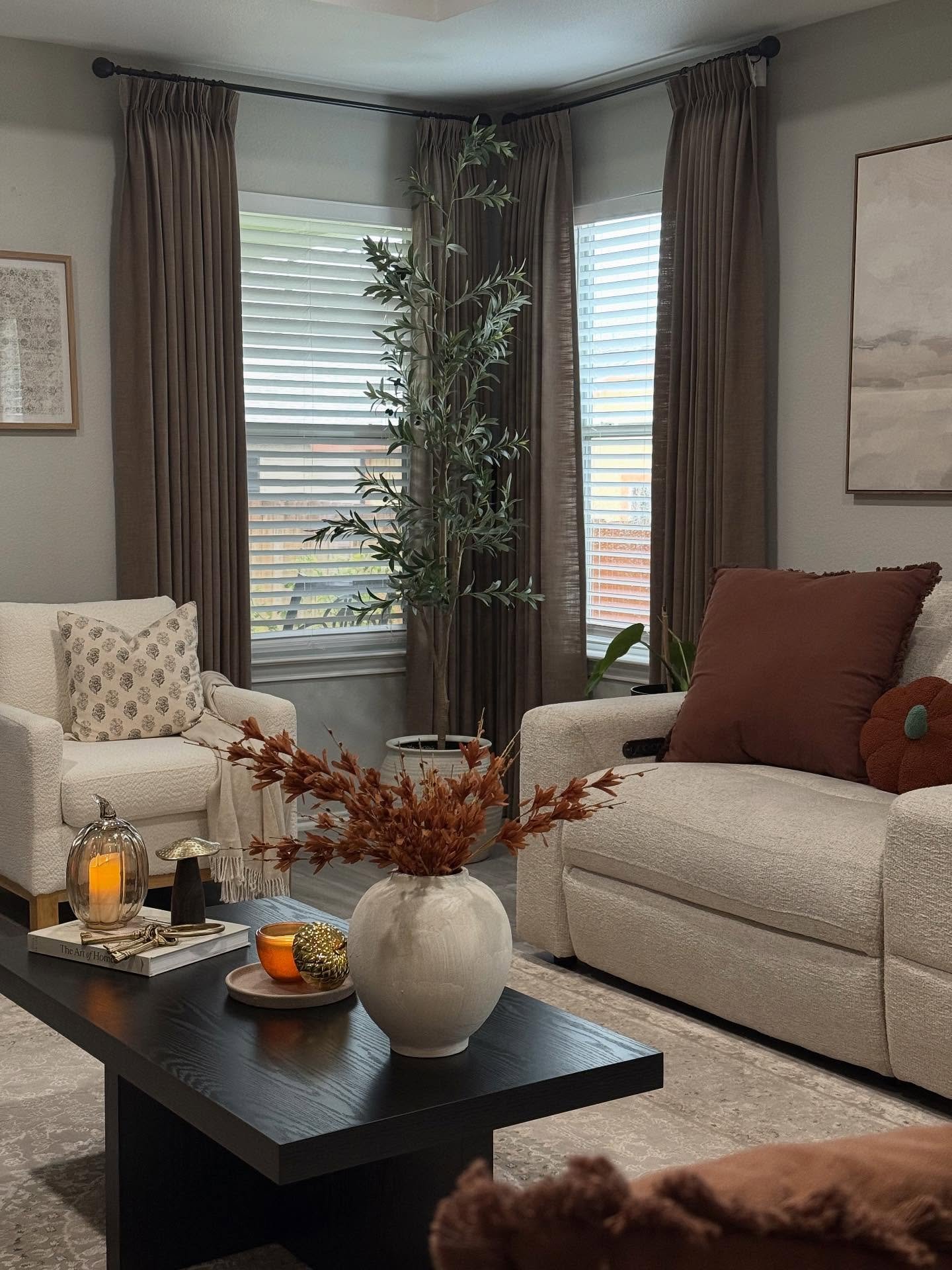 Living room with two light-colored armchairs, brown curtains, and a black coffee table with a white vase holding dried flowers.