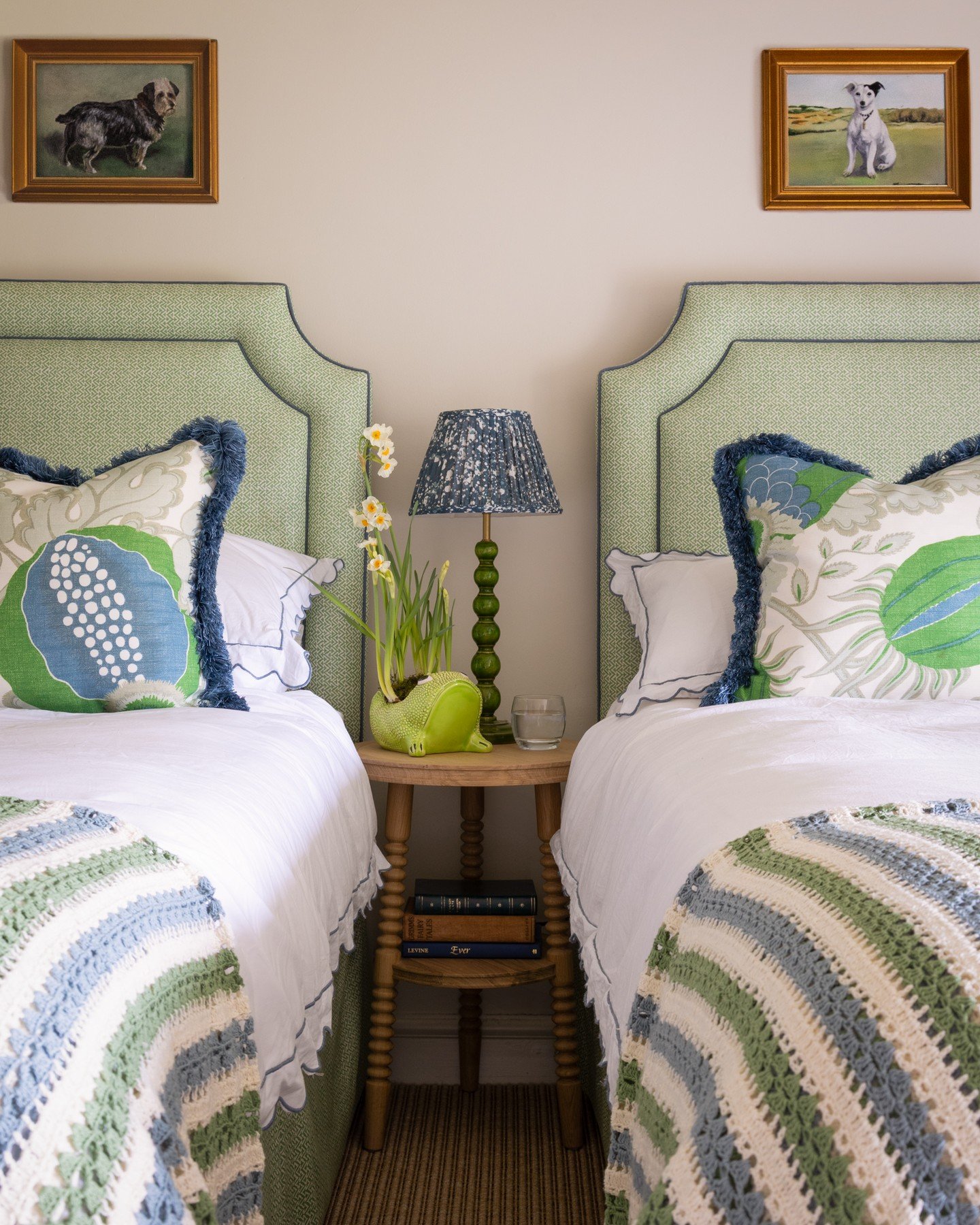 Twin bedroom with green upholstered headboards, striped crochet blankets, a wooden nightstand, and framed dog portraits.
