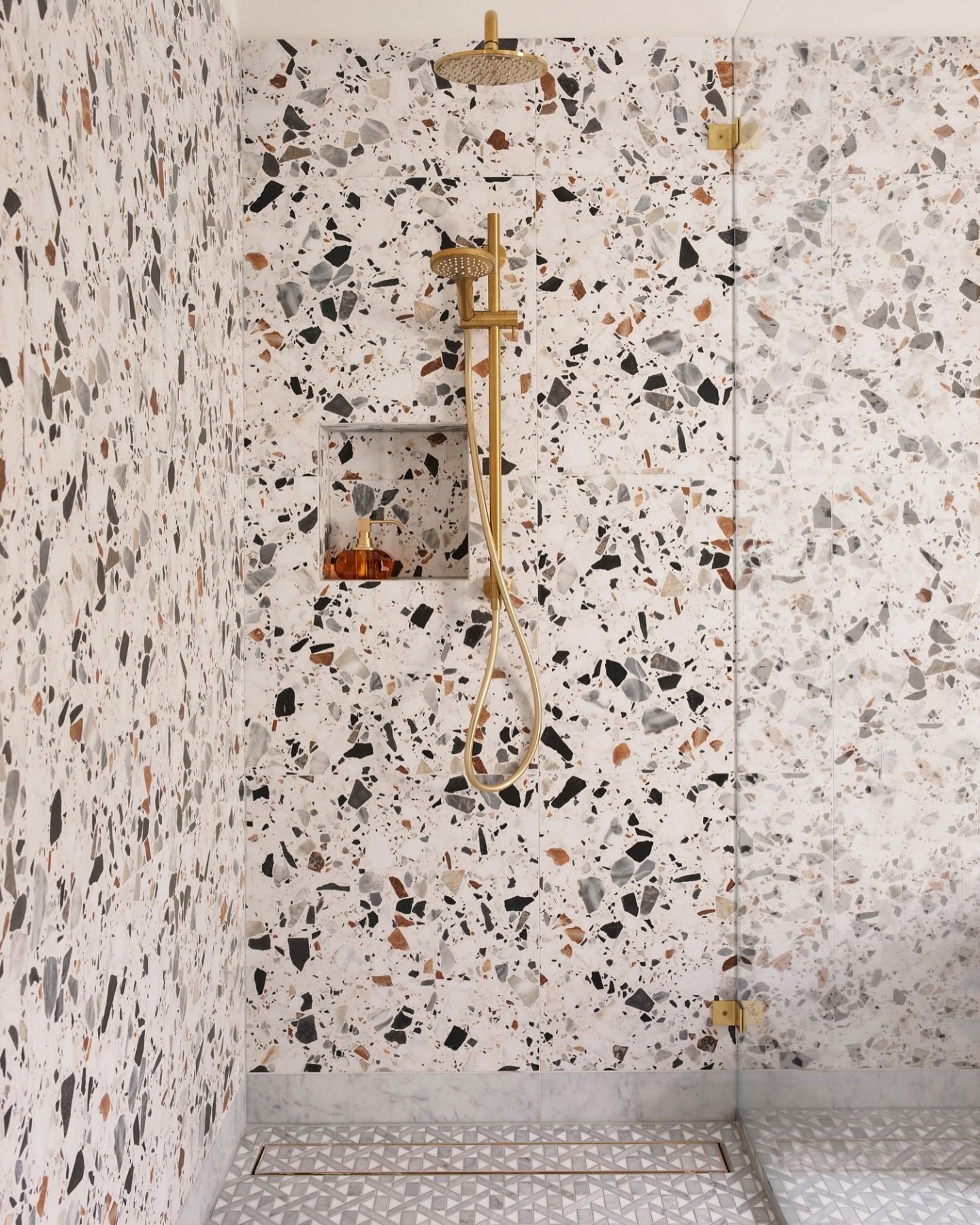 A walk-in shower with terrazzo-style tiles in a mosaic of white, black, gray, and warm brown flecks, with a smaller geometric pattern on the floor and brass fixtures.