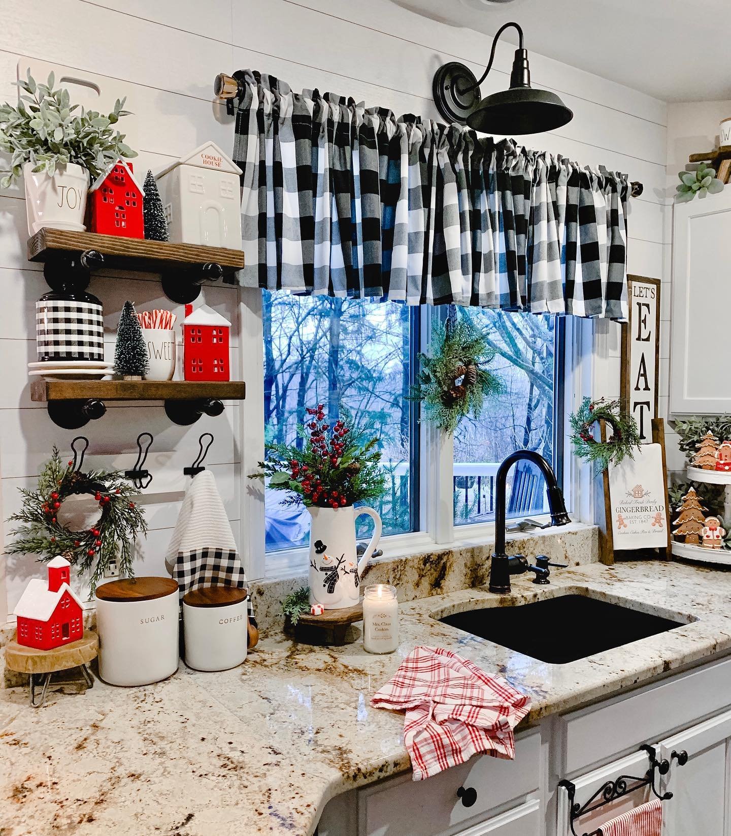 A festive farmhouse kitchen sink area decorated with a black and white buffalo check valance and red and black accents. Open pipe shelving holds miniature red house ceramics and Christmas trees, while small wreaths hang on the window.