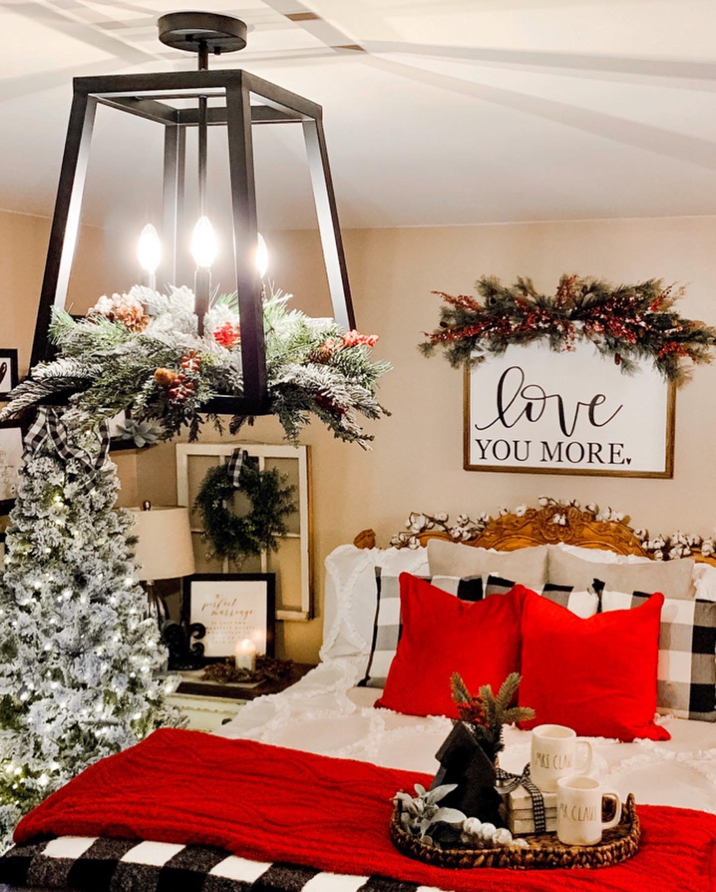 A cozy bedroom featuring a dramatic black lantern chandelier decorated with a flocked garland, a bed with red and black buffalo check pillows and a chunky red blanket, and a wall sign that reads "love you more."