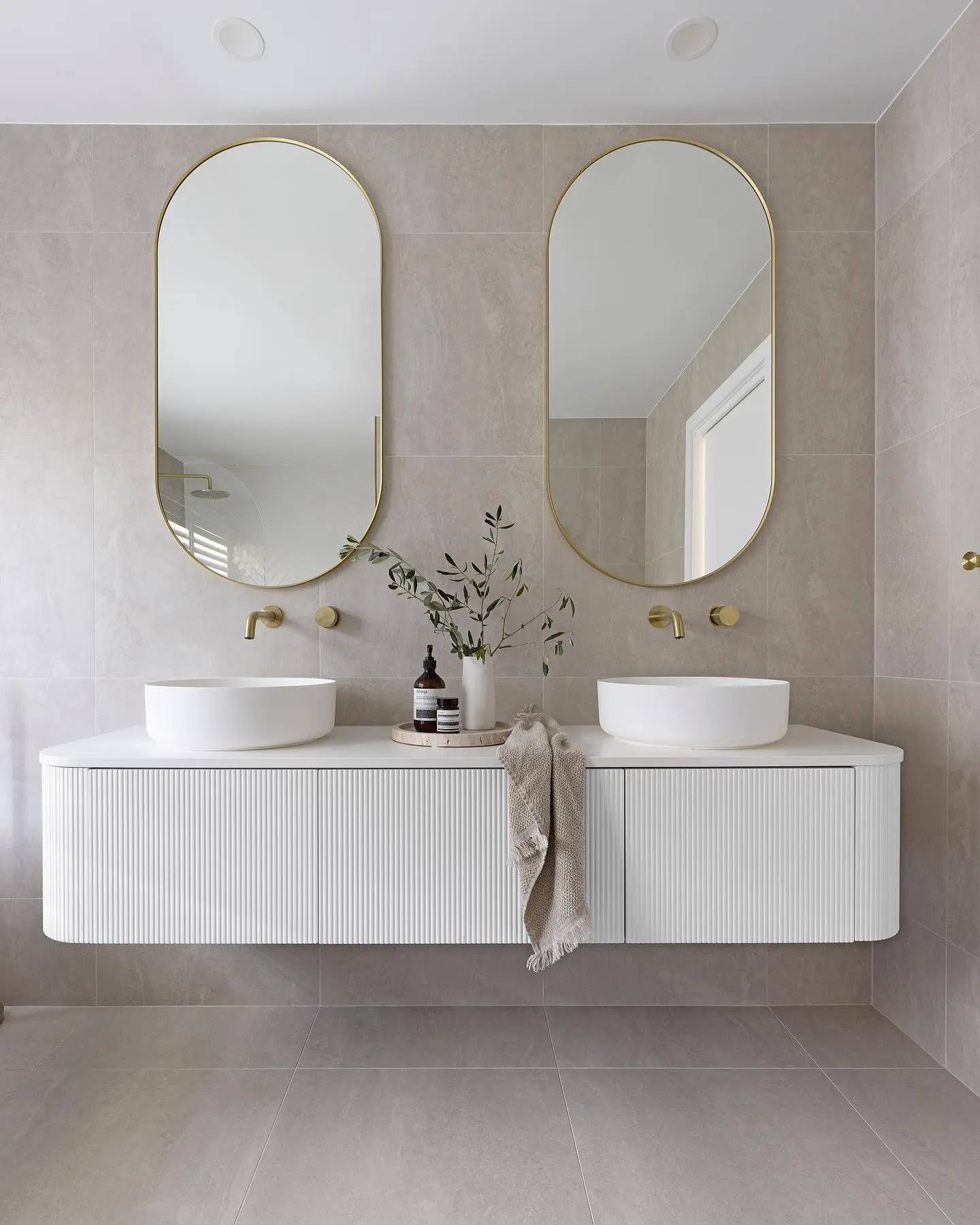 A minimalist bathroom with two identical tall, arched gold-framed mirrors over a white double vanity.