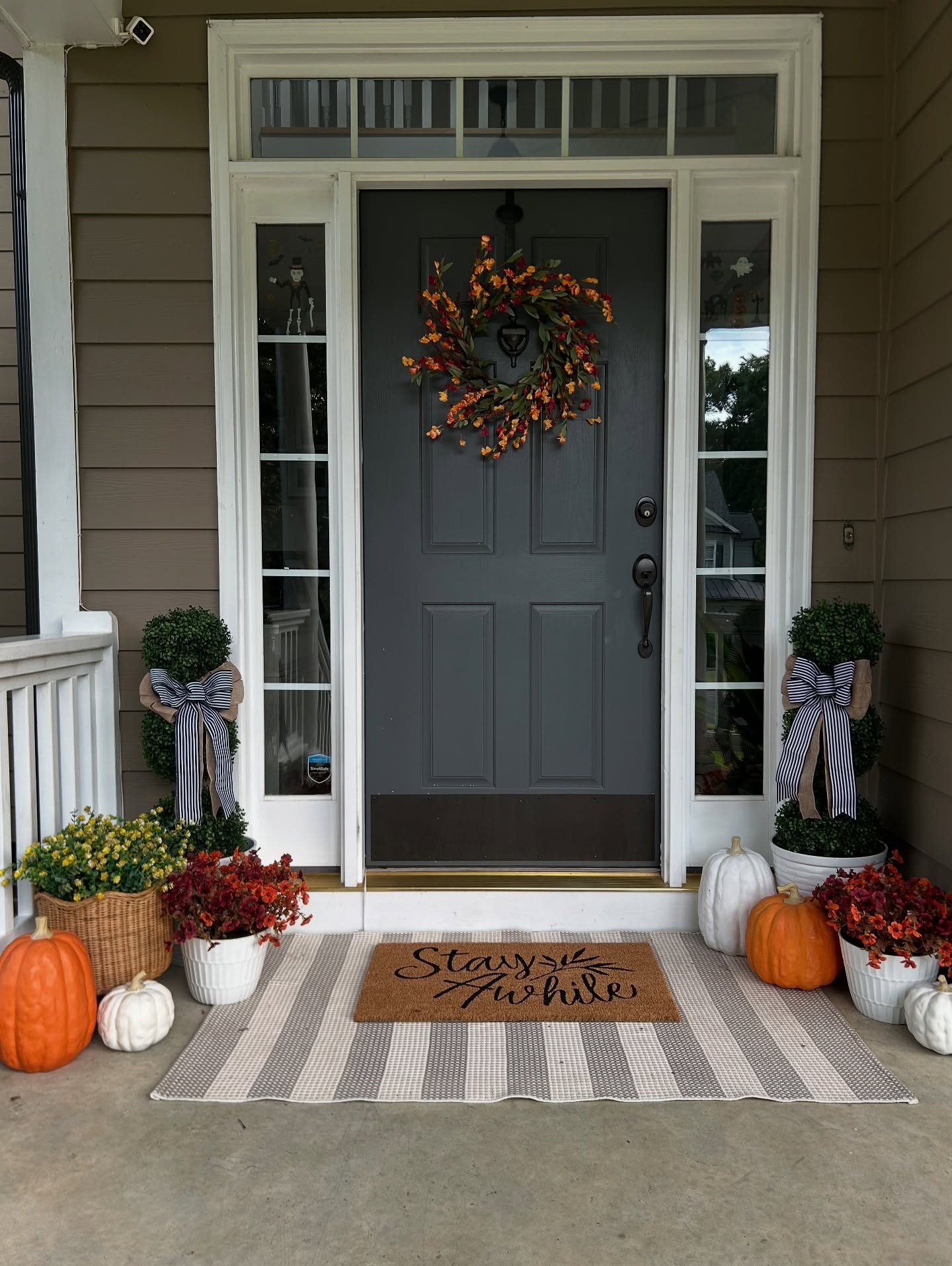 A symmetrical fall porch with a gray door, decorated with white pumpkins, orange pumpkins, planters of mums, and boxwood topiaries with striped bows.