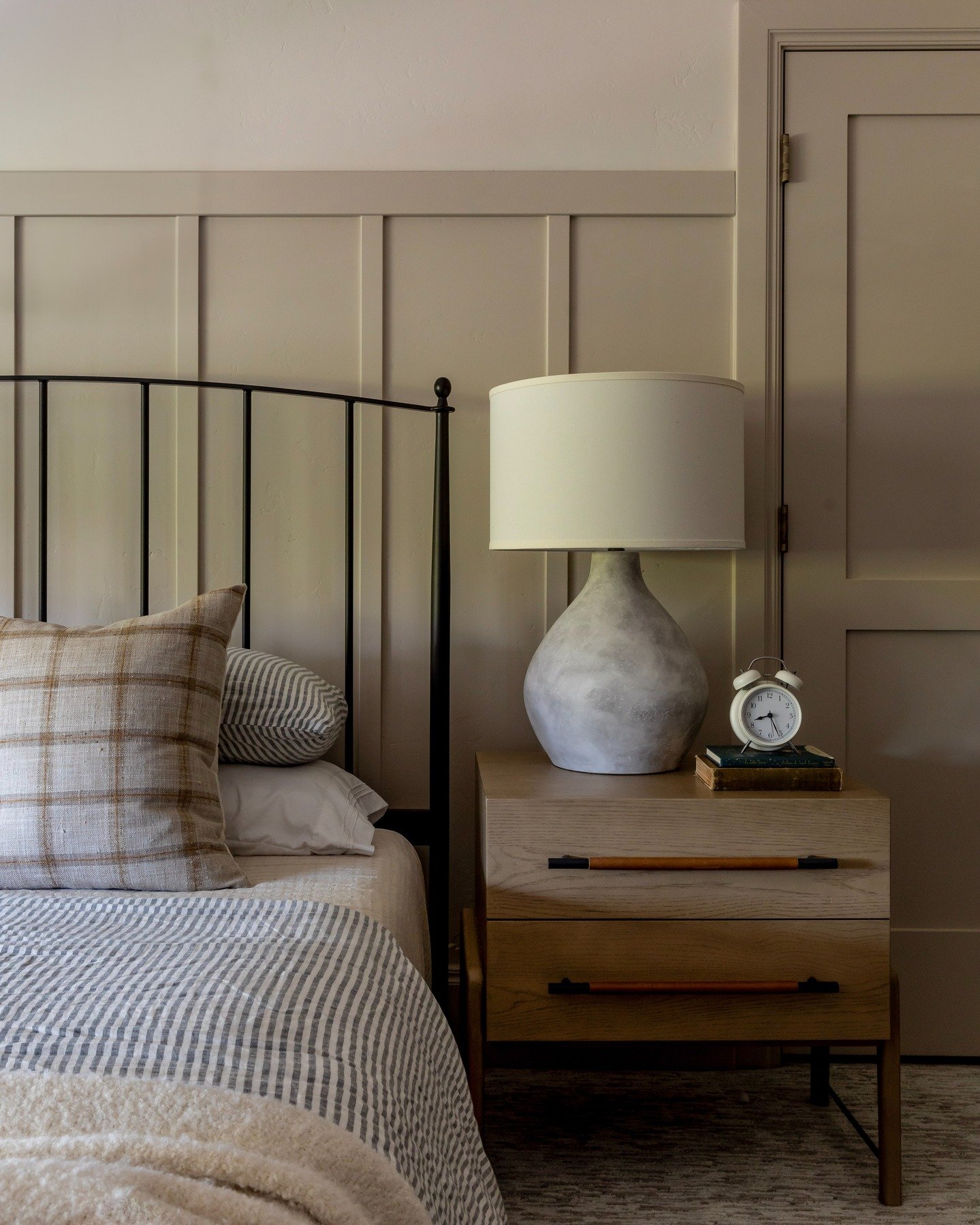 A light wood nightstand with two drawers featuring black and brown leather strap pulls. On the top sits a large, textured gray ceramic lamp and a small vintage-style alarm clock.