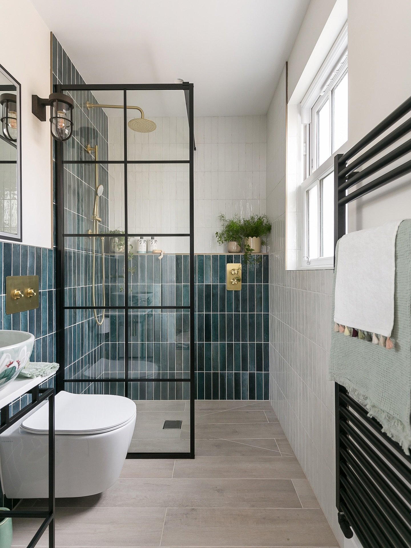 A narrow bathroom with a Crittall-style black metal shower screen, featuring walls tiled halfway in glossy teal subway tiles and the rest in light gray tiles.