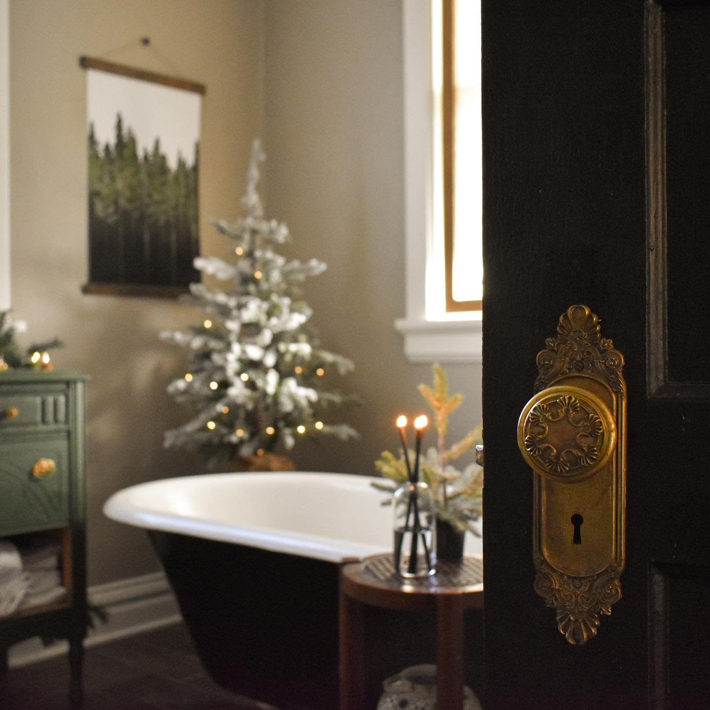 A dark, moody bathroom with a black clawfoot tub, featuring a flocked and lit Christmas tree and a forest wall hanging.