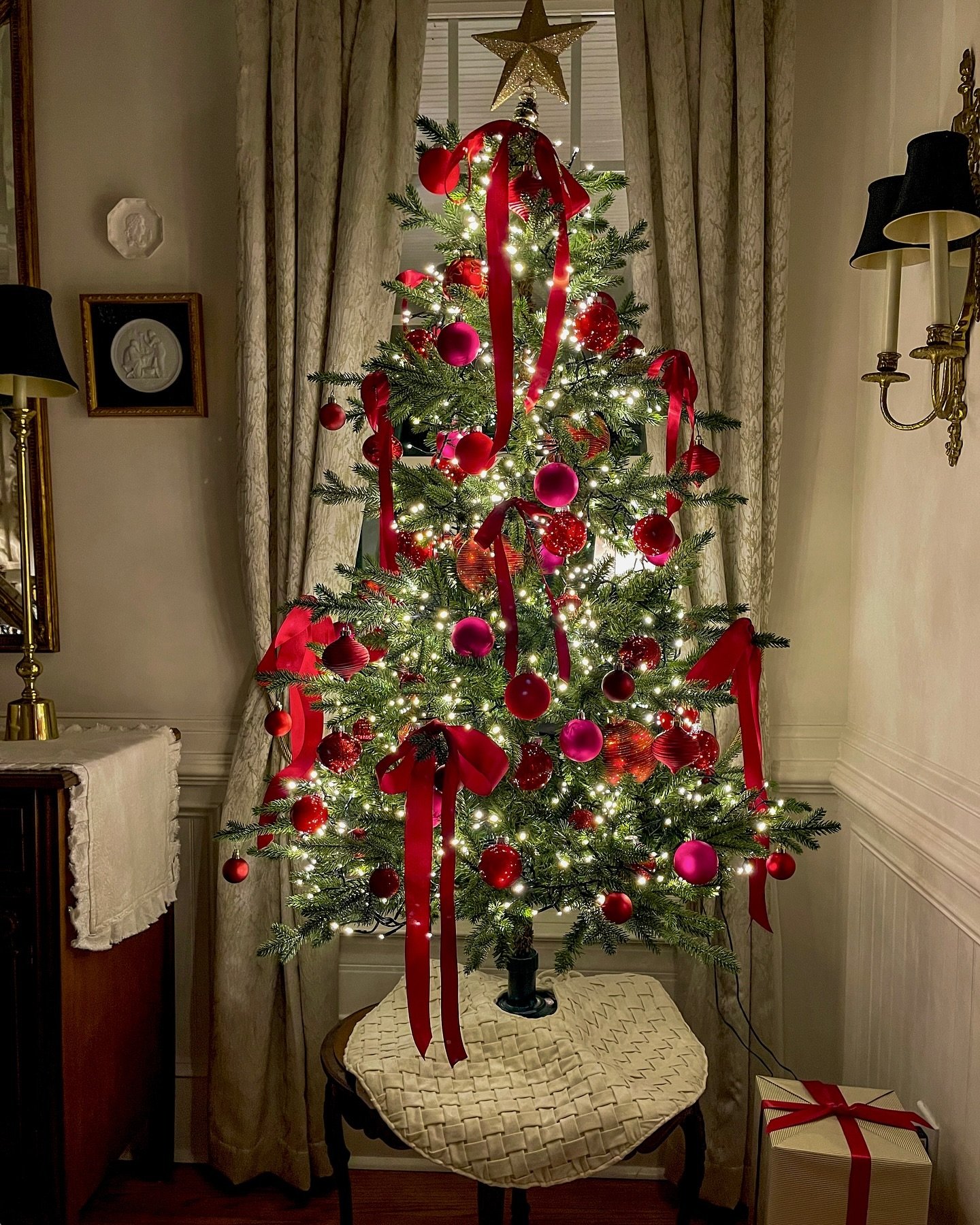 A slim Christmas tree decorated with deep red and vibrant magenta ornaments, featuring cascading red velvet ribbons, set on a small side table.