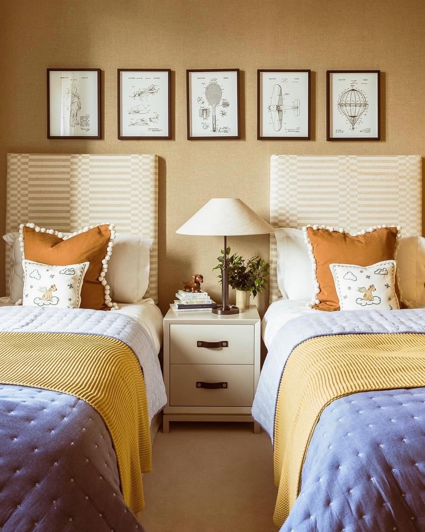 Twin bedroom with beige patterned headboards, yellow and lavender bedding, a white nightstand, and framed vintage patent artwork.