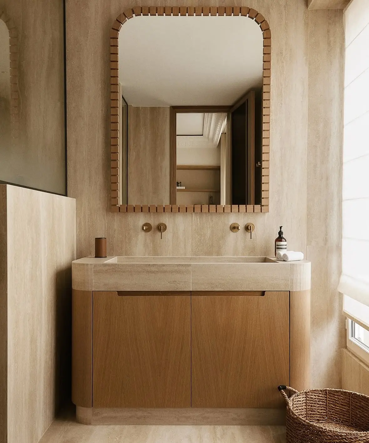 A double vanity with a light wood cabinet and a stone countertop with two integrated sinks. It is paired with a matching wood-framed rectangular mirror with rounded corners, against a travertine-tiled wall.