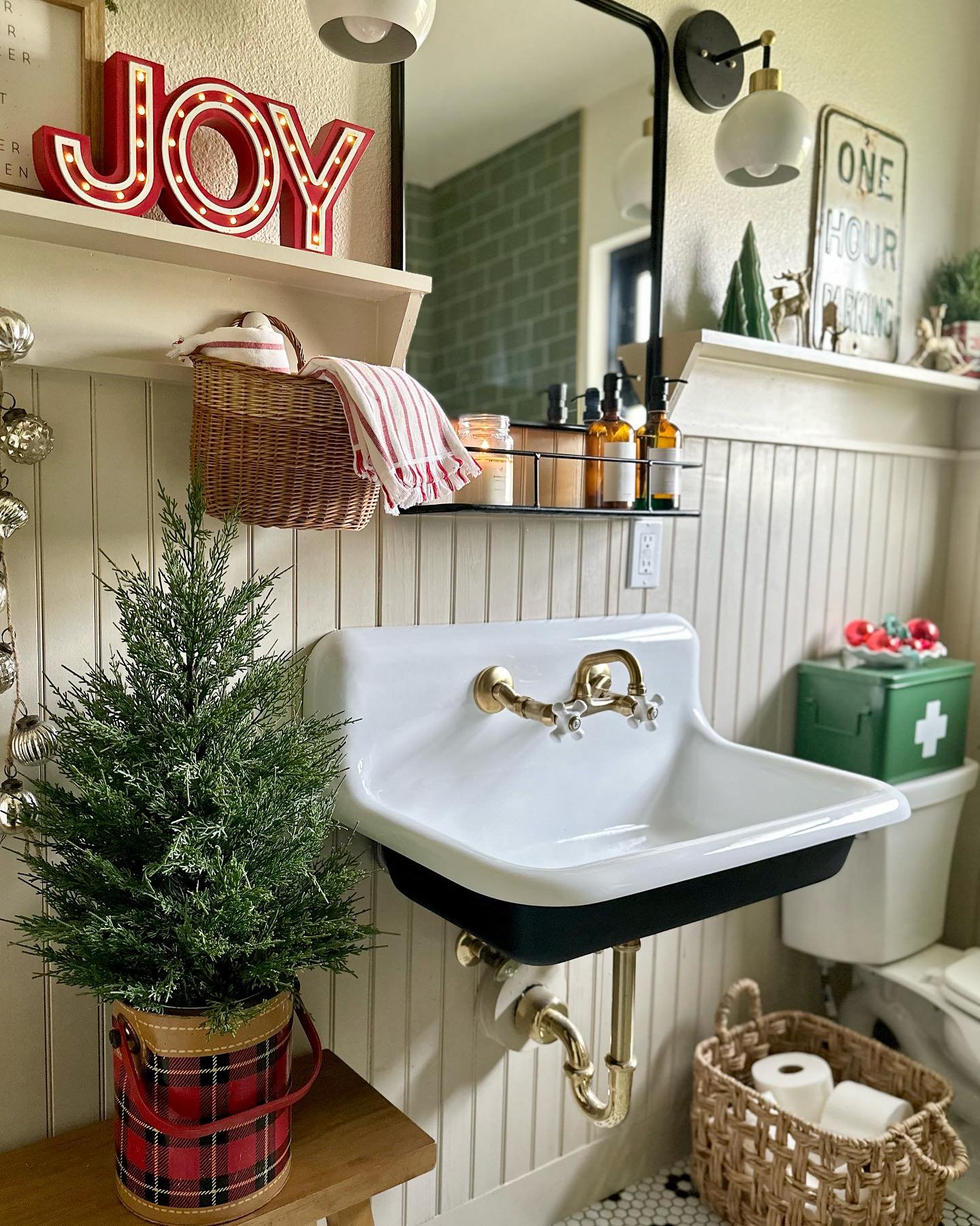 A vintage-style sink in a beadboard bathroom decorated for Christmas with a plaid tree bucket, a red "JOY" marquee sign, and a green storage box.