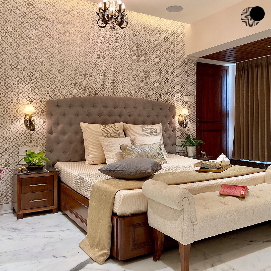 A cozy bedroom with a large tufted headboard, patterned wallpaper, and rich, dark wood furniture.