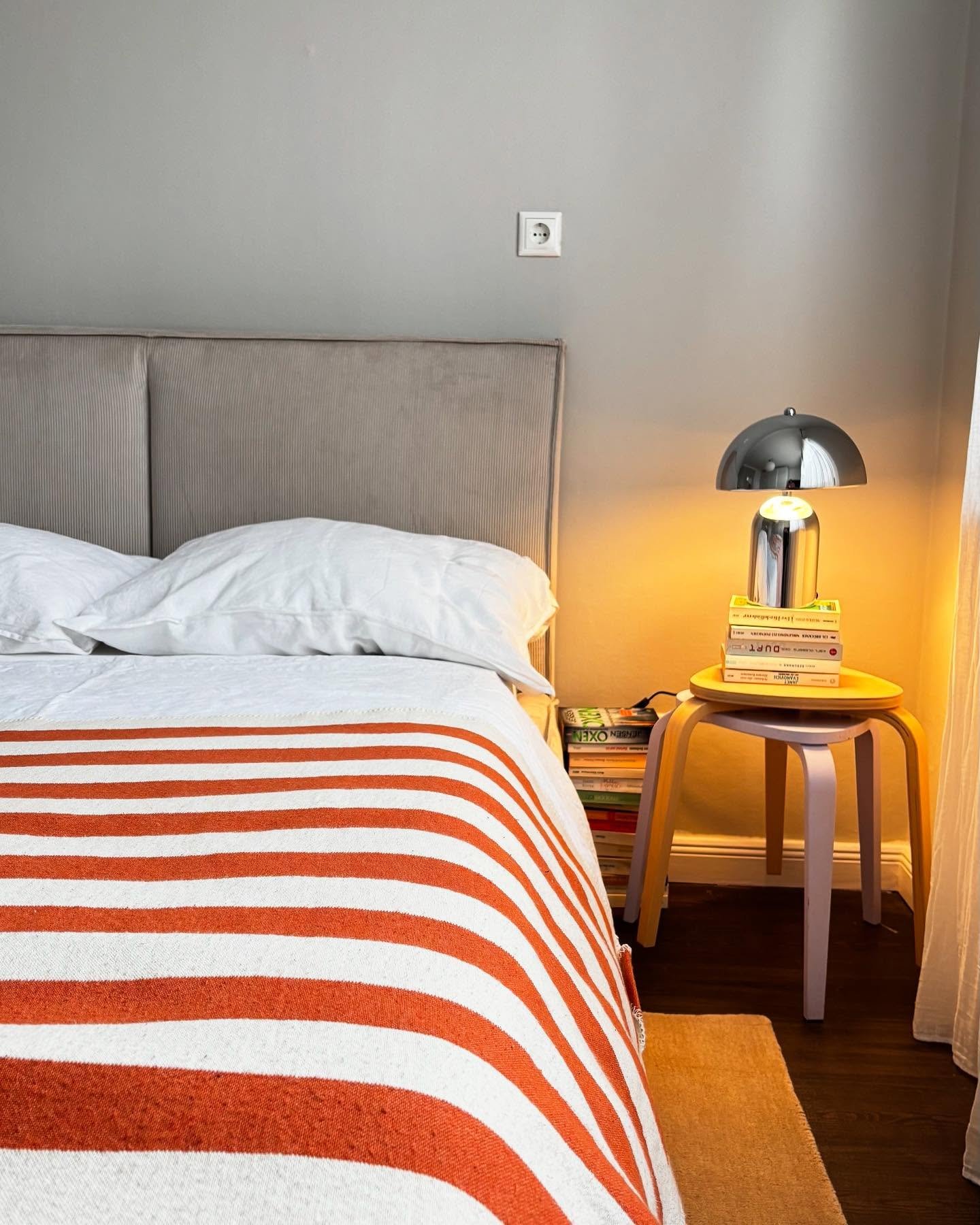 A small, mushroom-shaped table lamp with a chrome finish sits on a yellow and purple wooden stool used as a nightstand.