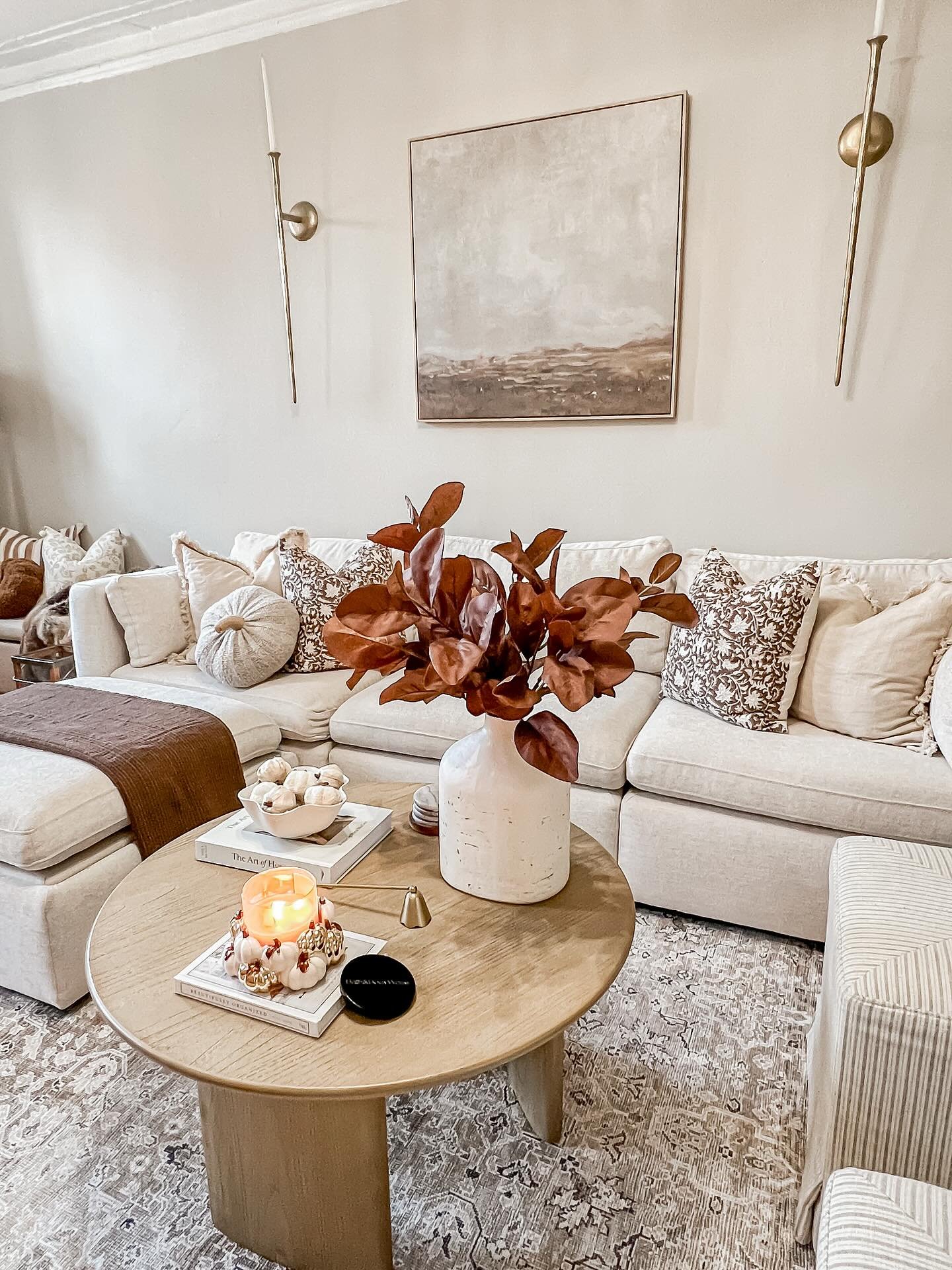 Living room with a large, light-colored sectional sofa, a round wooden coffee table, and a vase with large russet-colored leaves.