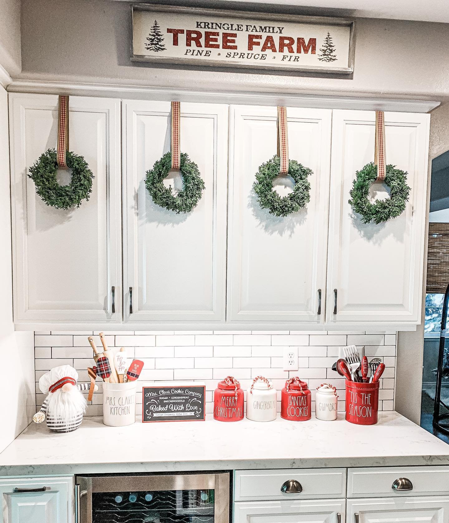A white kitchen featuring four small green wreaths hung symmetrically on upper cabinet doors with red and white checkered ribbons. The counter displays a set of red and white labeled holiday canisters and a "Kringle Family Tree Farm" sign overhead.