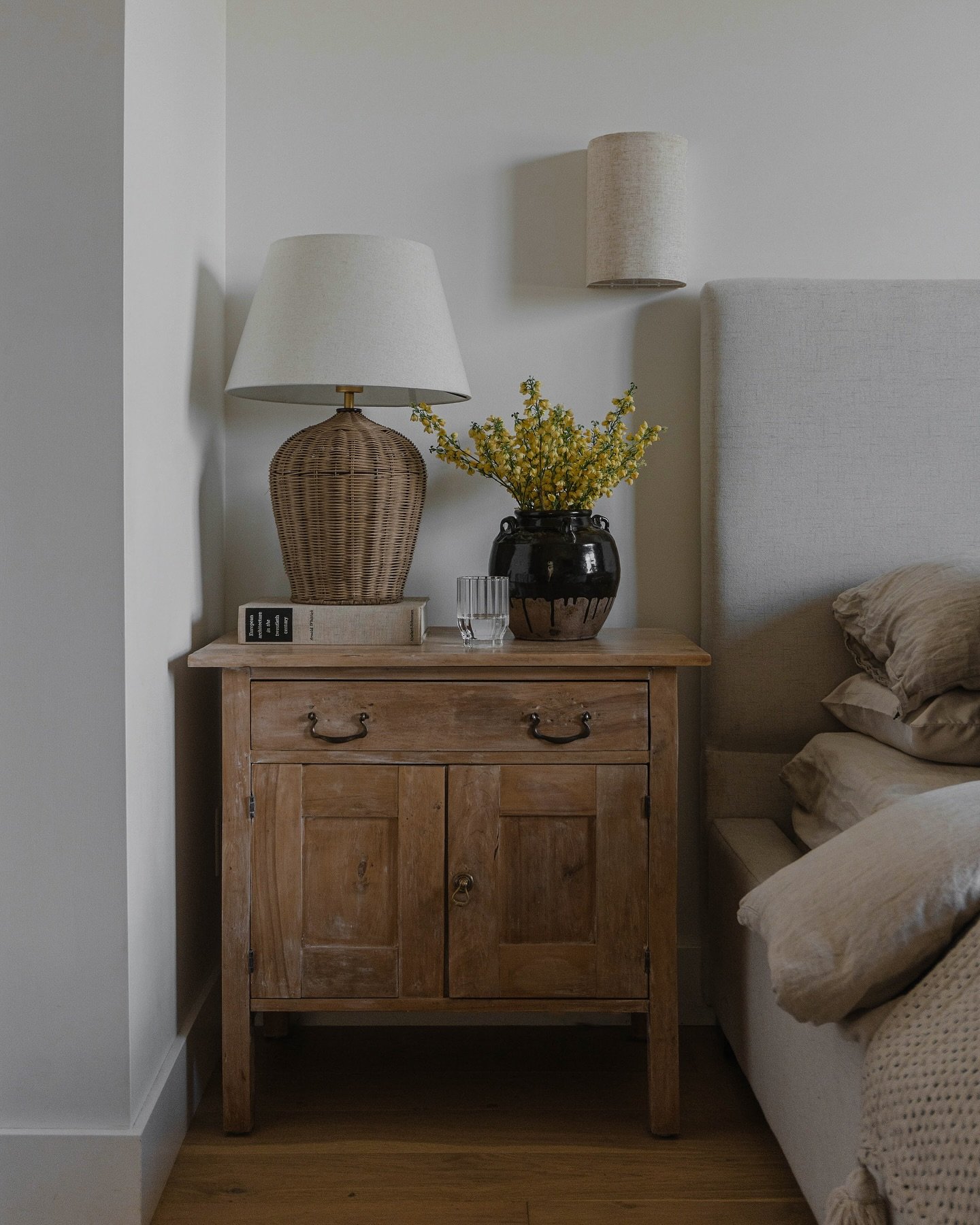 An old, rustic wooden nightstand with one drawer and two lower cabinet doors with aged metal ring pulls. The top holds a stack of books, a dark ceramic vase with yellow flowers, a glass of water, and a large lamp with a woven wicker base.