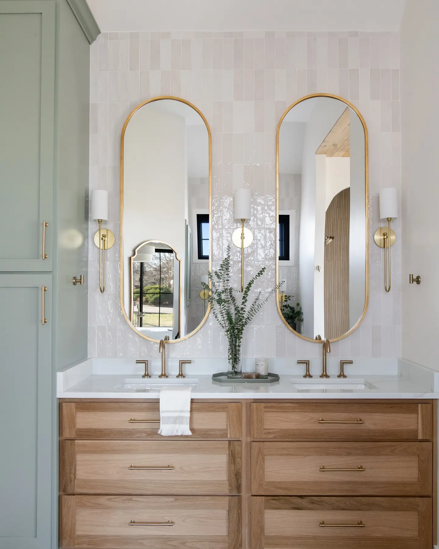 A bright bathroom with a double wood vanity and two identical tall, arched gold-framed mirrors over each sink.