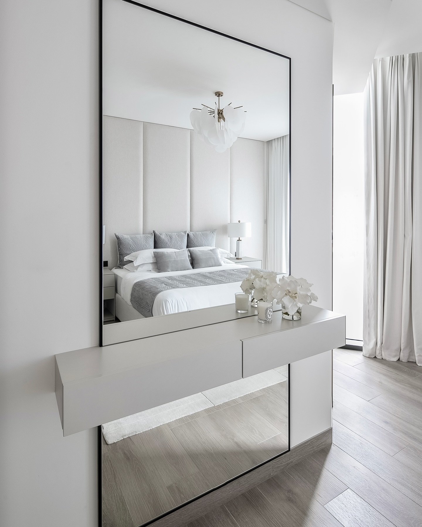 A large, floor-to-ceiling mirror with a thin black frame is integrated with a floating white vanity in a modern bedroom, reflecting the bed.