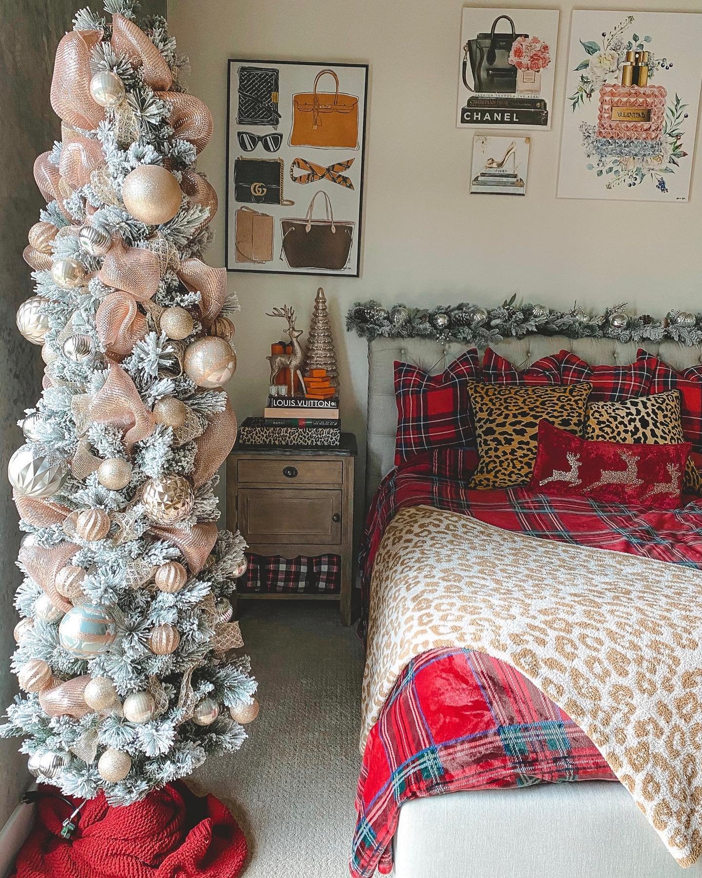 A stylish bedroom featuring a slim, heavily flocked Christmas tree decorated with rose gold ribbon and ornaments, a bed with red and black plaid bedding, and contrasting leopard print pillows and throw.
