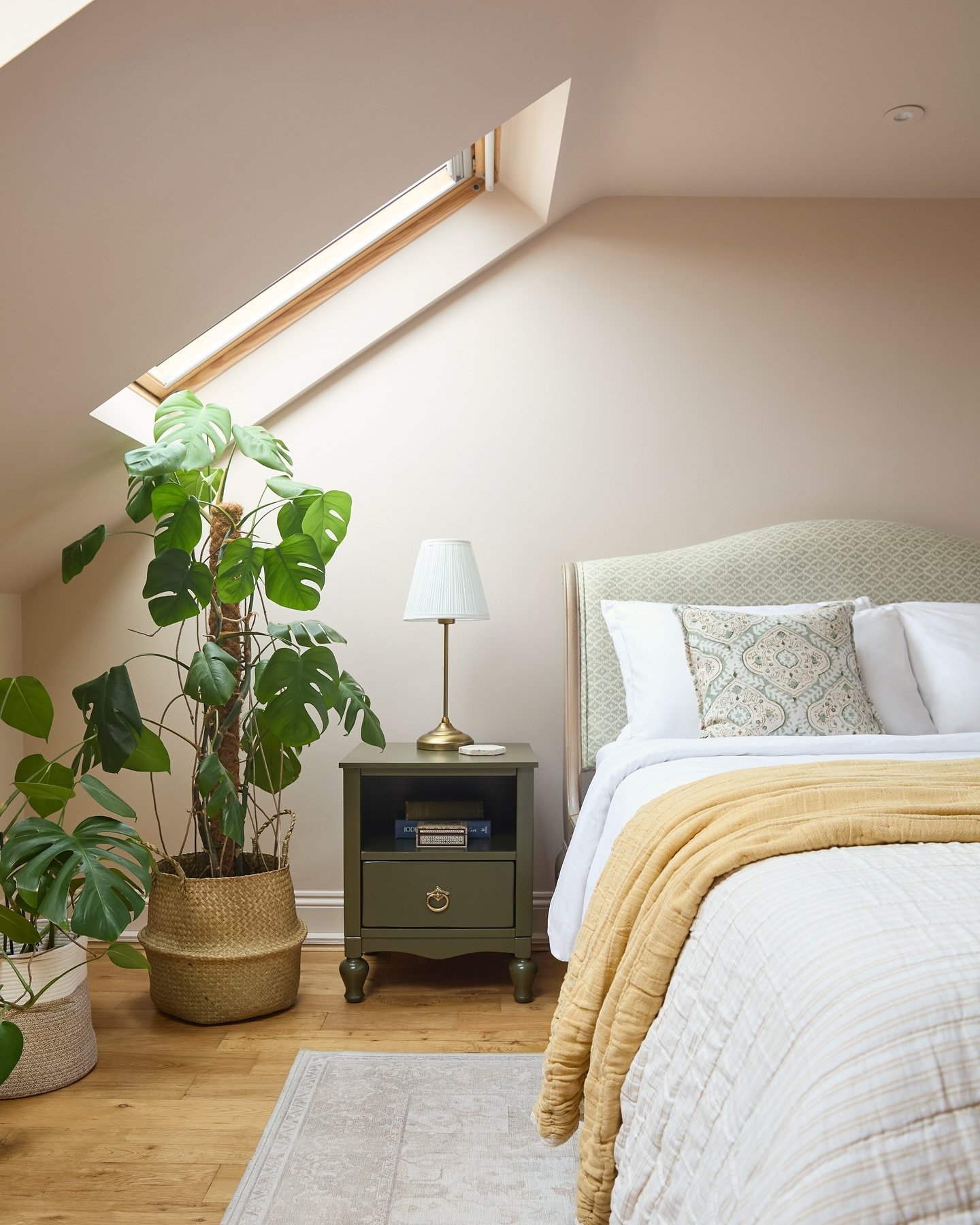 A natural and bright loft bedroom with a large plant, a light-colored arched headboard, and an olive-green nightstand.