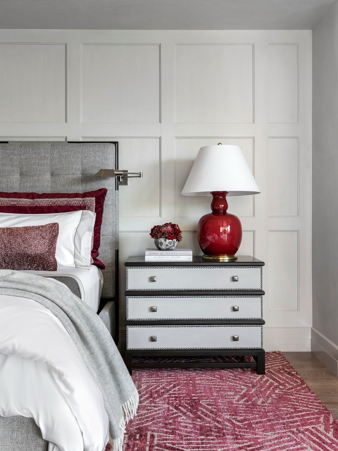 A dark wood nightstand with three light-colored drawers featuring a unique stitched detail along the edges and metallic stud-like pulls. A large, bright red ceramic lamp with a white shade and a small vase of dark red flowers sits on top.