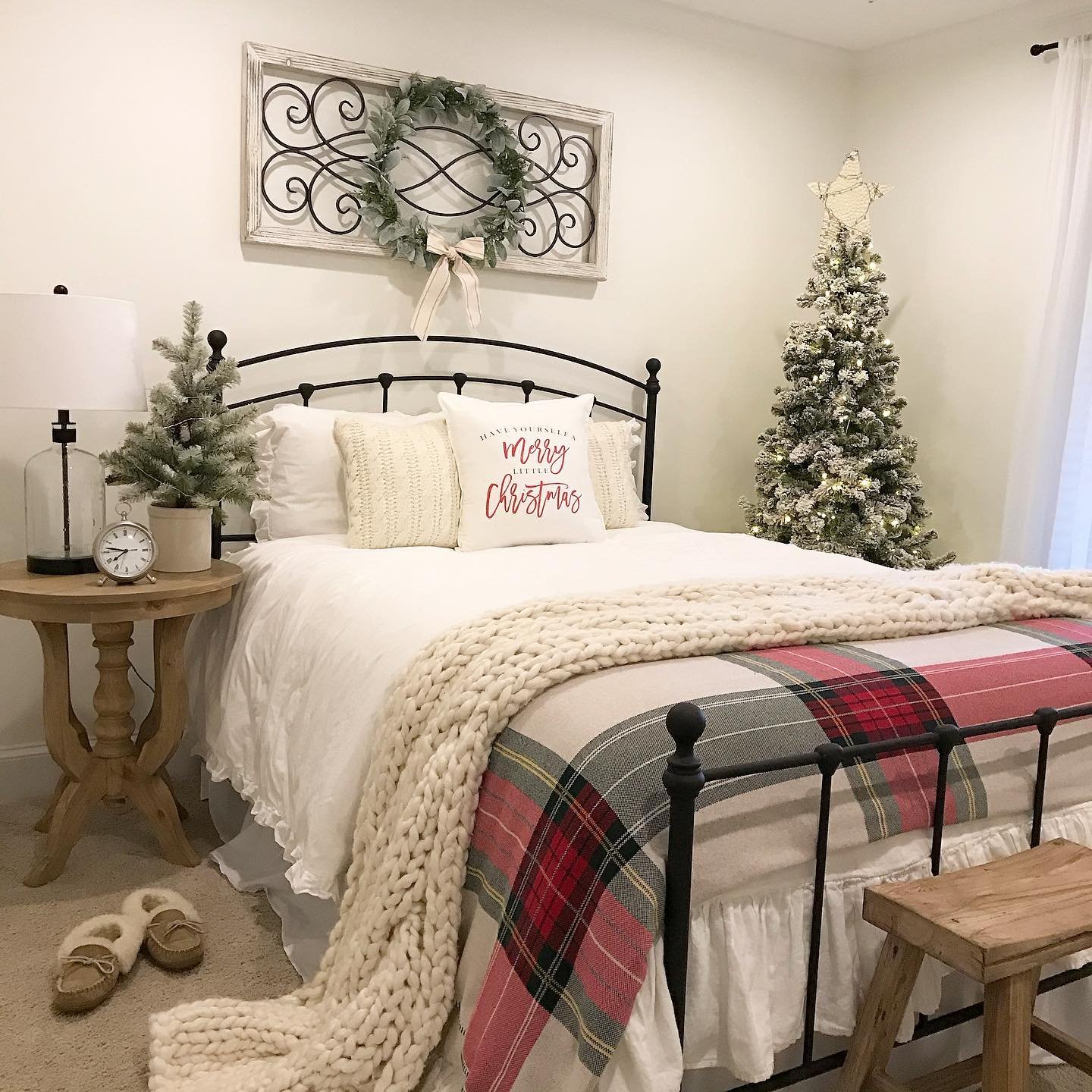 A bright bedroom with a black metal bed frame, white bedding, a red and gray plaid blanket, a chunky knit throw, and a flocked Christmas tree with a star topper.