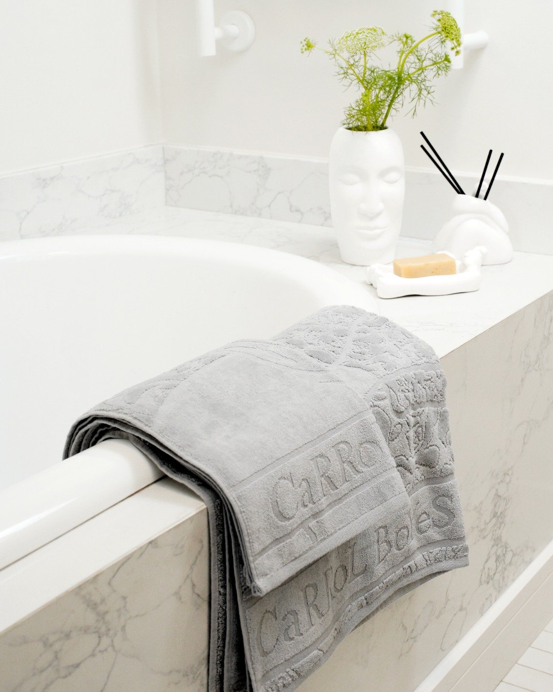 Dove grey monogrammed towel draped over a white bathtub with a marble surround and white sculptural accessories.