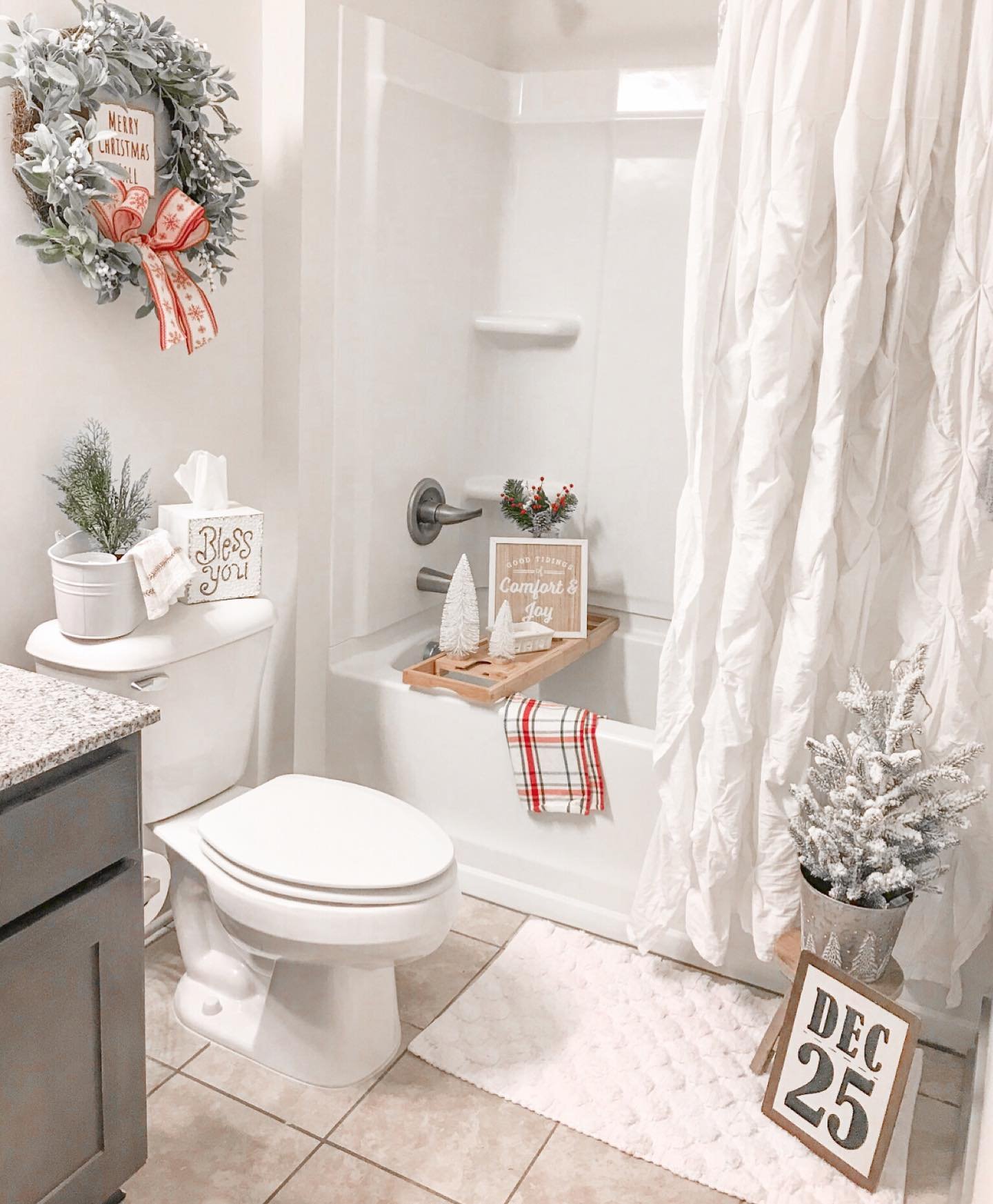 A white and grey bathroom decorated for Christmas with a flocked wreath, miniature white trees on a bath tray, and a flocked tree on a "DEC 25" sign.