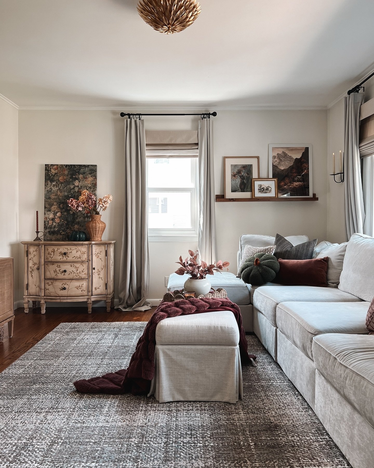 A vintage-style living room with a light gray sectional, an antique painted chest, and a gallery wall with mountain art.