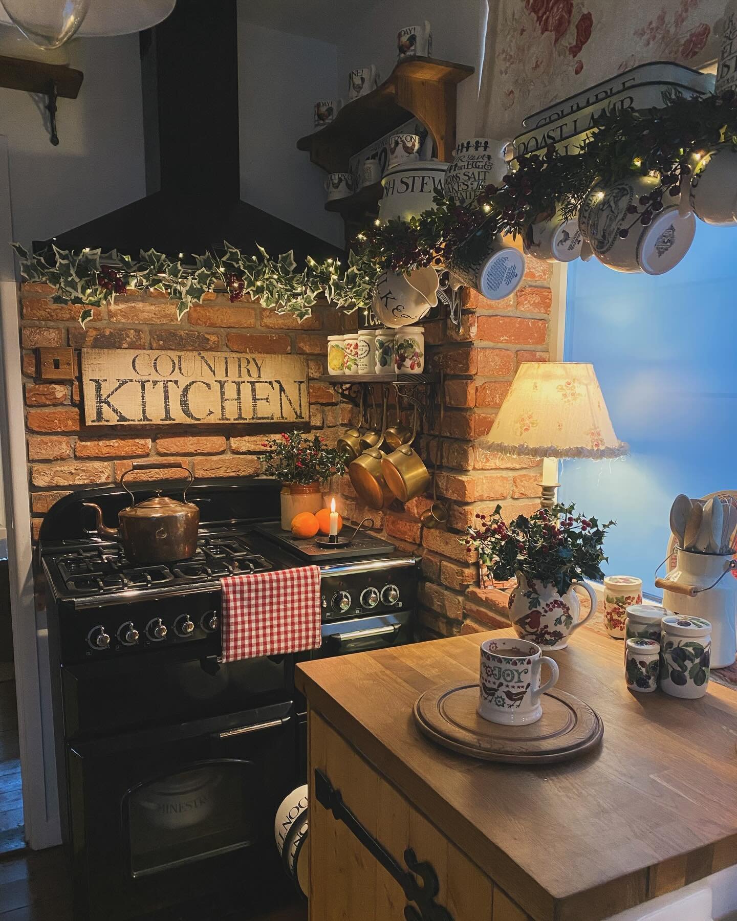 A warm, traditional kitchen featuring exposed brick, a black range, and wood countertops. Festive decor includes an ivy garland with string lights, copper pots, and vintage Christmas mugs.
