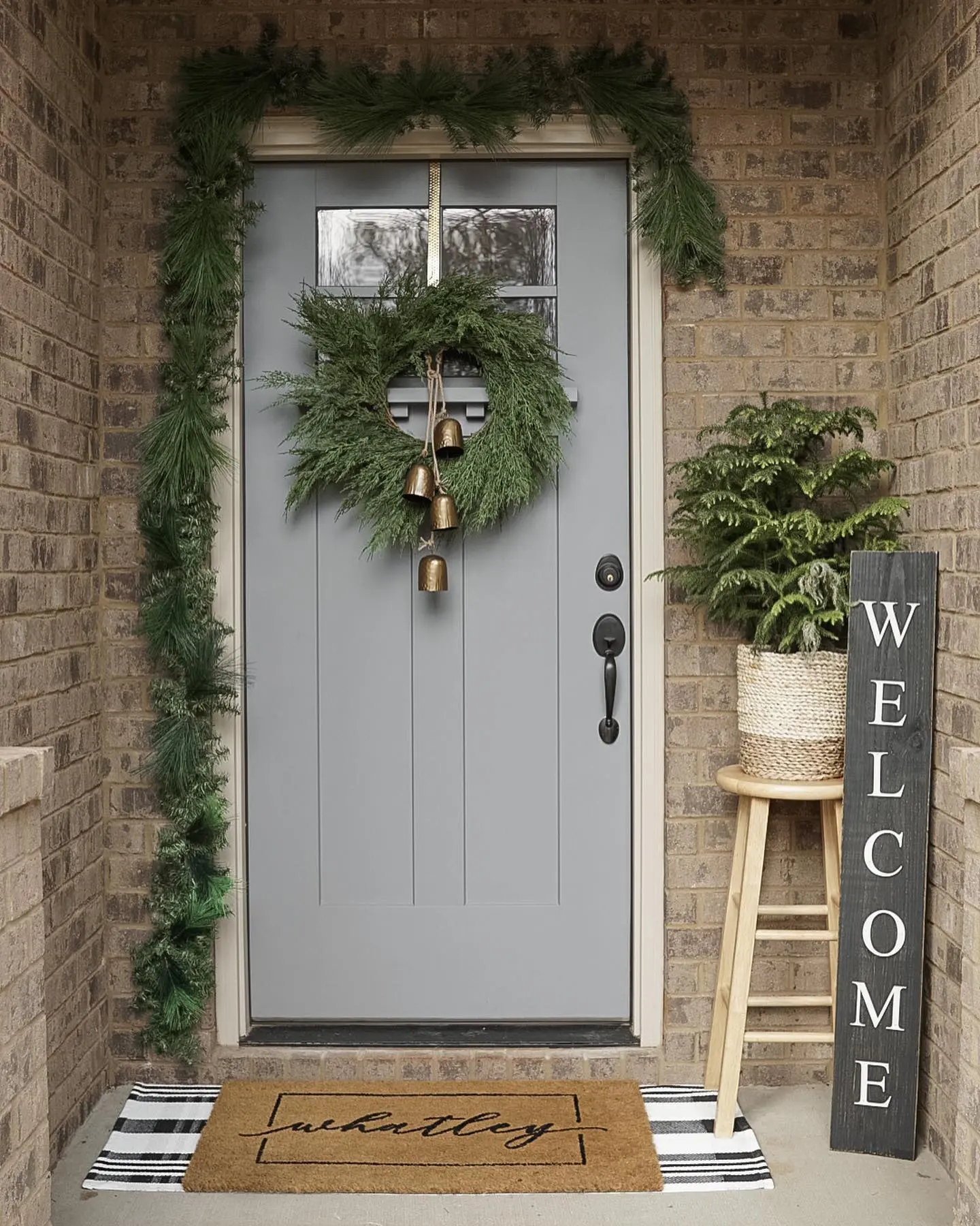 A contemporary Christmas porch with a light gray door, featuring a simple cedar wreath hung with brass bells, sparse evergreen garland, and a black and white striped layered doormat.