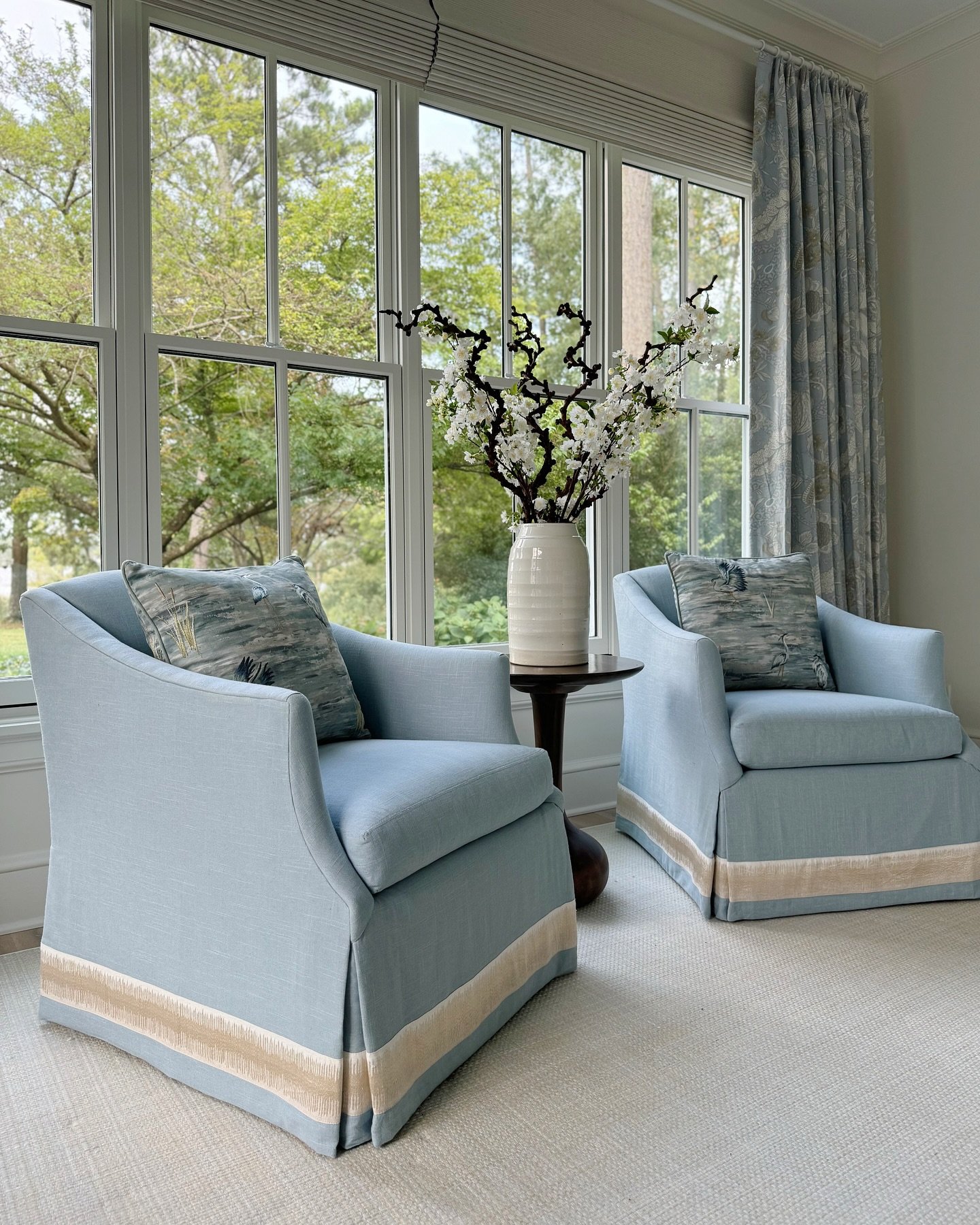 A pair of light blue skirted armchairs with a contrasting fringe border sits facing each other in a sunlit room.
