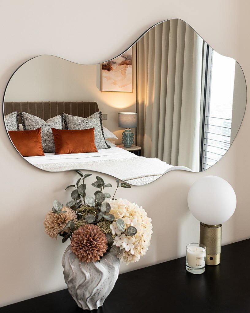  large, frameless mirror with an abstract, wavy shape hangs on a wall, reflecting a bed with brown and orange pillows.