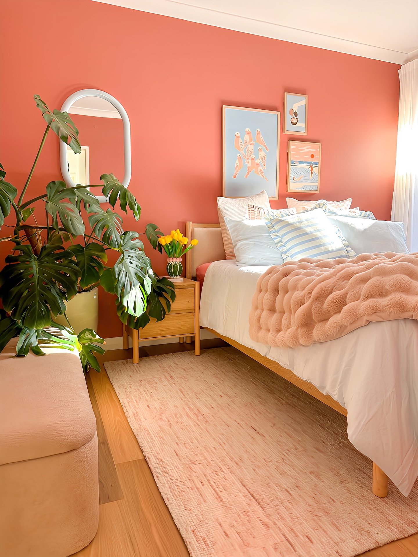 A vibrant bedroom with a peach accent wall, a bed with a blush-colored faux fur throw, a large monstera plant, and a gallery wall with various art prints.